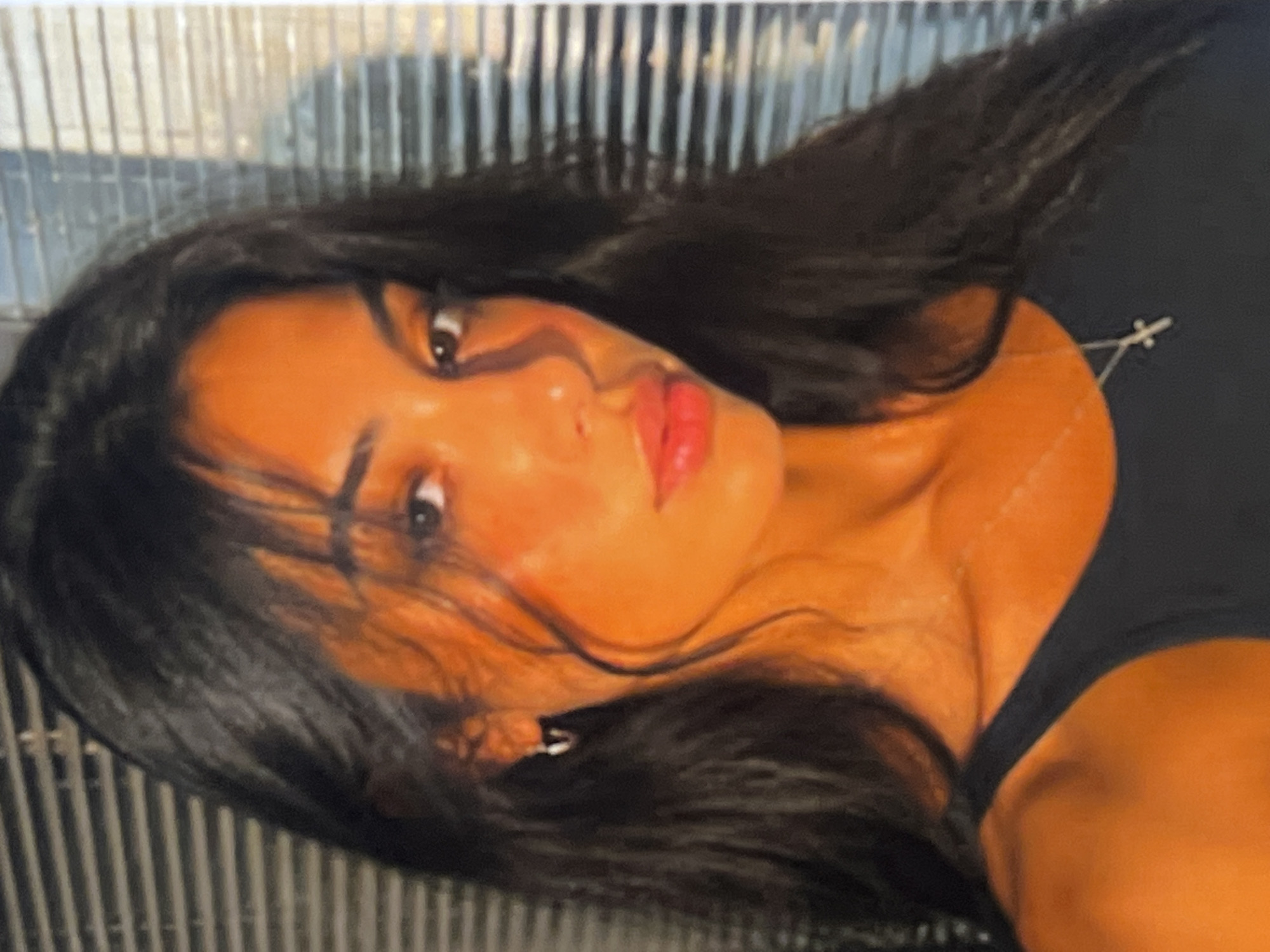 An extreme close-up, medium shot captures a young woman with dark, long, wavy hair, her head tilted slightly to her left, looking directly at the camera. Her skin is illuminated by warm, orange light, creating strong highlights and shadows across her face. She is wearing a black tank top and a small, silver cross necklace. A small silver hoop earring is visible in her left ear. The background consists of horizontal blinds with a textured, possibly brick, pattern behind them, suggesting an indoor environment. The lighting appears to be artificial or from a strong, directional light source.