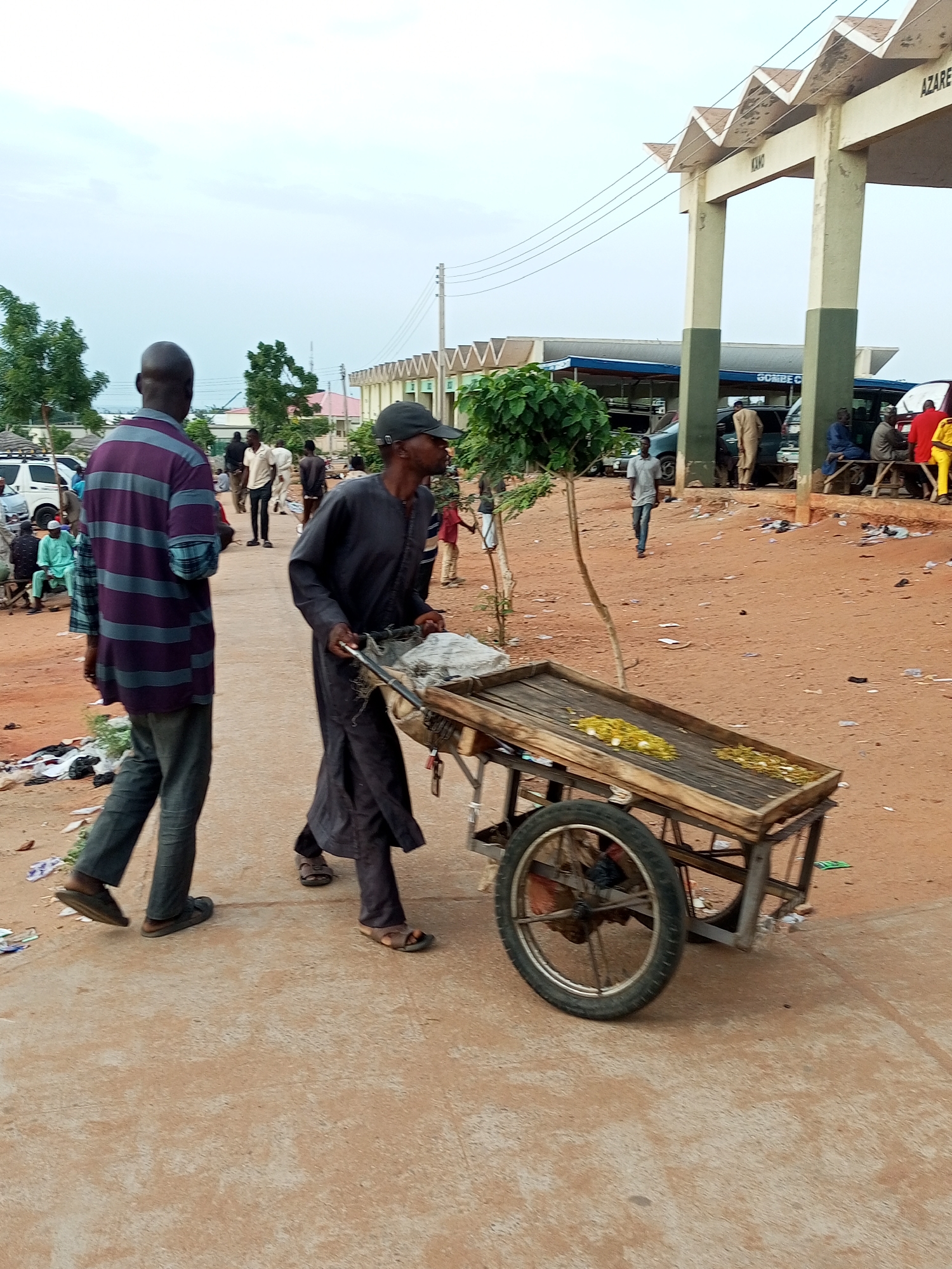 The image shows a street scene in Gombe, Nigeria, under an overcast sky. In the foreground, a man in a dark robe is pushing a wooden cart with a single wheel. The cart contains some white bags and what appears to be yellow food items. To the left of the cart pusher, a man in a purple and grey striped shirt and dark pants walks away from the camera. Behind them, other individuals are visible, some standing and some sitting. Several vehicles, including a white van and other cars, are parked in the background. A large, architectural structure with zig-zagging decorative elements and supporting pillars, possibly an entrance or a covered market area, is visible on the right side of the image, with text that appears to say "KANO" and "AZARE". The ground is predominantly reddish-brown dirt, with scattered litter. Trees are visible on the left side of the image.