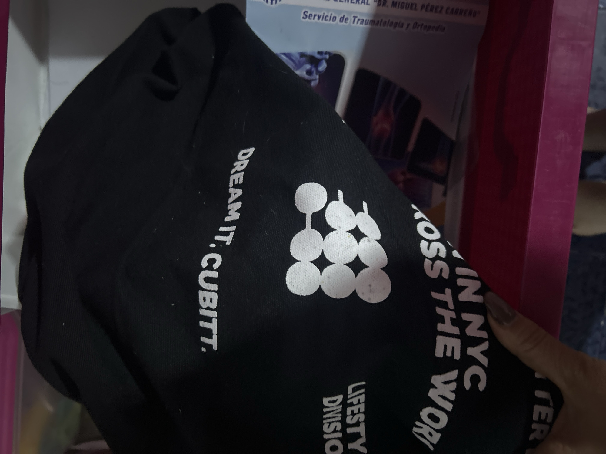 A close-up shot shows a black garment, likely a shirt or hoodie, with white text and a logo printed on it. The text reads "DREAM IT. CUBITT.", "LIFESTY DIVISION", and "IN NYC KOSS THE WOR". A hand with a manicured nail is holding a part of the garment. In the background, there is an out-of-focus box with text that reads "DR. MIGUEL PÉREZ CARREÑO" and "Servicio de Traumatologia y Ortopedia". The color of the box is pink and white.
The information provided does not state the city of San Juan de Los Morros, Venezuela.