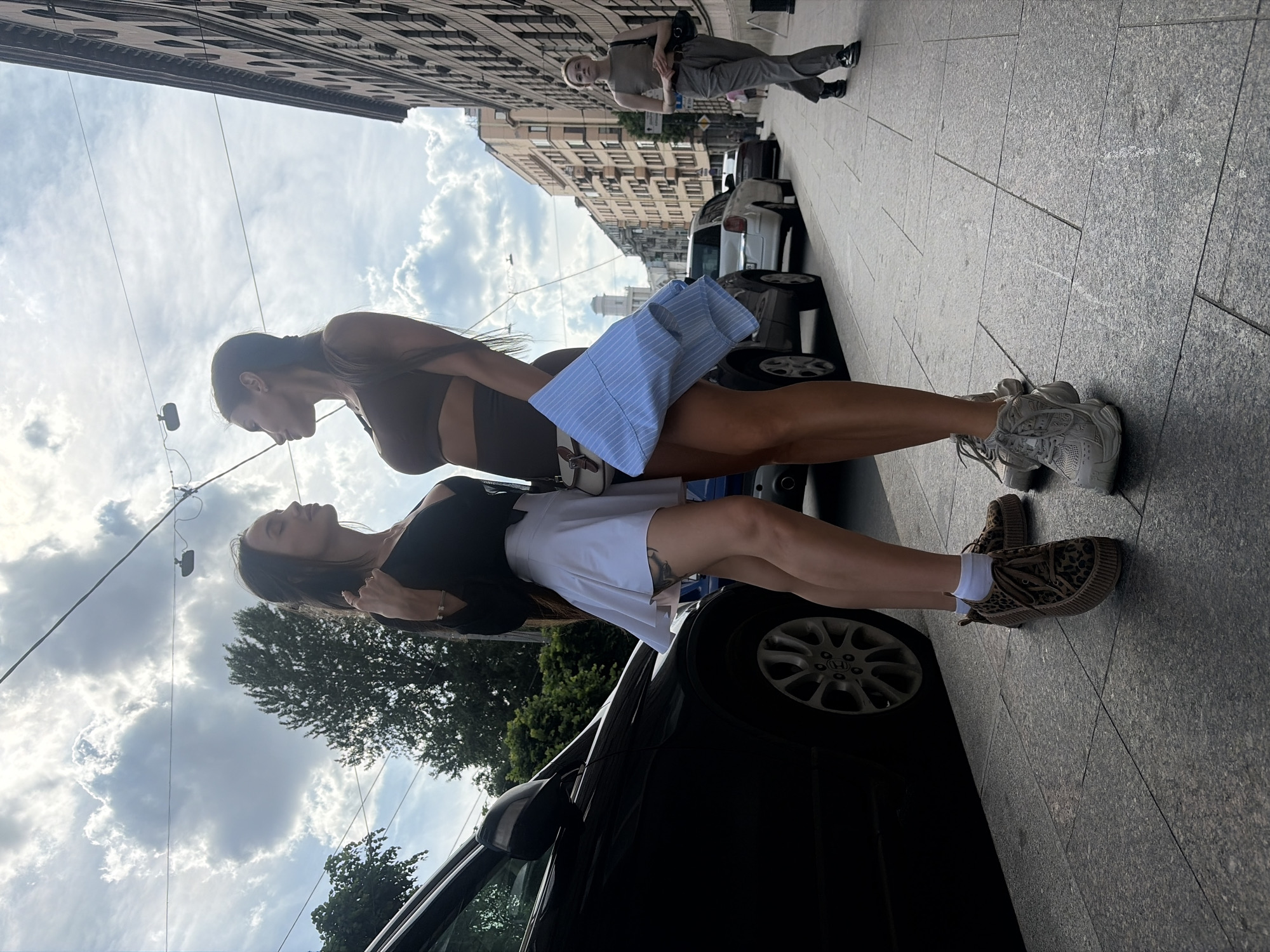 The image depicts an urban street scene in Saint Petersburg, Russia, likely along Izmailovsky Prospekt or a nearby street, with the Trinity Cathedral (Troitsky Sobor) visible in the background.
In the foreground, two women are positioned facing each other on a granite-paved sidewalk under bright, partly cloudy skies. The woman on the left, with long dark hair, wears a black long-sleeved top, a white pleated mini-skirt, white socks, and leopard-print platform sneakers. A tattoo is visible on her left thigh. She has a bracelet on her right wrist, and her right hand is raised towards her face. The woman on the right, also with long dark hair styled in a braid, wears a dark brown athletic crop top and matching shorts, along with white chunky sneakers. She carries a white shoulder bag and holds a folded light blue striped shirt in her left hand. Both women are looking directly at each other, standing in close proximity.
To the left, a portion of a black car or SUV is parked parallel to the sidewalk. In the mid-ground, a large tree is visible. Overhead wires, possibly for power or public transport, crisscross the sky. On the right side of the image, a row of multi-story, classical-style buildings lines the street. Further down the street on the right sidewalk, another woman is walking away from the viewer. She wears a light brown sleeveless top, grey pleated trousers with a black belt, and black shoes, carrying a black shoulder bag. Multiple cars are parked along the street on the right. In the distant background, the distinctive light-colored dome and tower structure of the Trinity Cathedral are visible above the buildings.