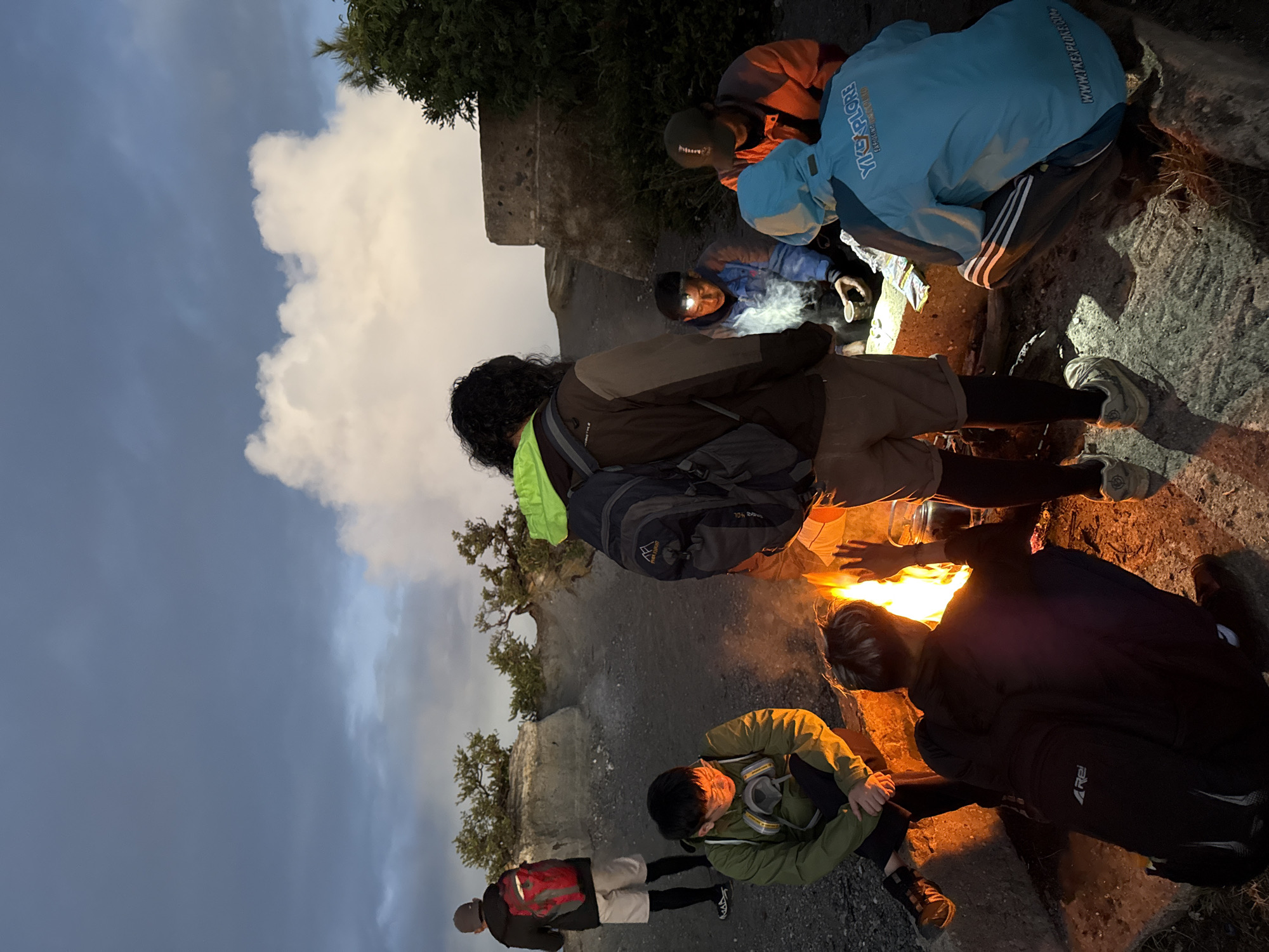 The image shows a group of people gathered around a fire, likely at dusk or dawn given the sky conditions. The location is Unknown, Indonesia.
The people are dressed in various hiking or outdoor clothing, suggesting they may be camping or trekking. They appear to be warming themselves by the fire or perhaps cooking. One person has a headlamp on. There are backpacks and other camping gear visible.
The terrain appears rocky and somewhat elevated, with some sparse vegetation. There is a large white cloud in the sky. The fire is a focal point, emitting a bright orange glow.