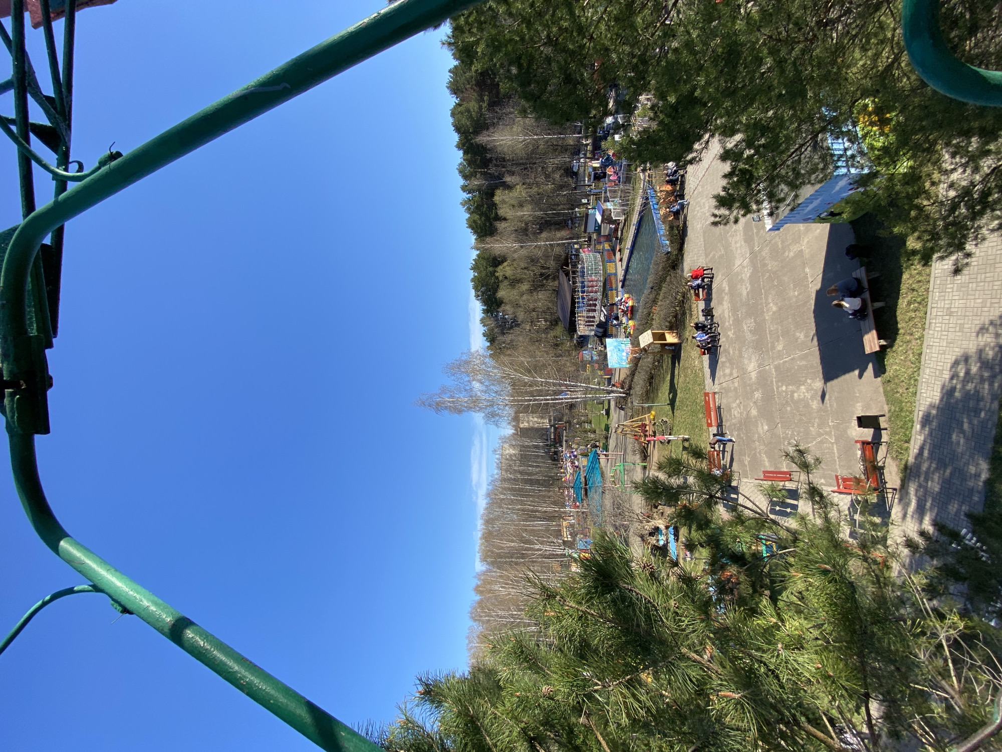The image shows a park in the city of Chaikovsky, Russia. The view is from an elevated position, possibly a Ferris wheel or similar structure. The park has a playground, benches, trees, and a pool. People are sitting on the benches and walking around the park. The weather appears to be sunny.