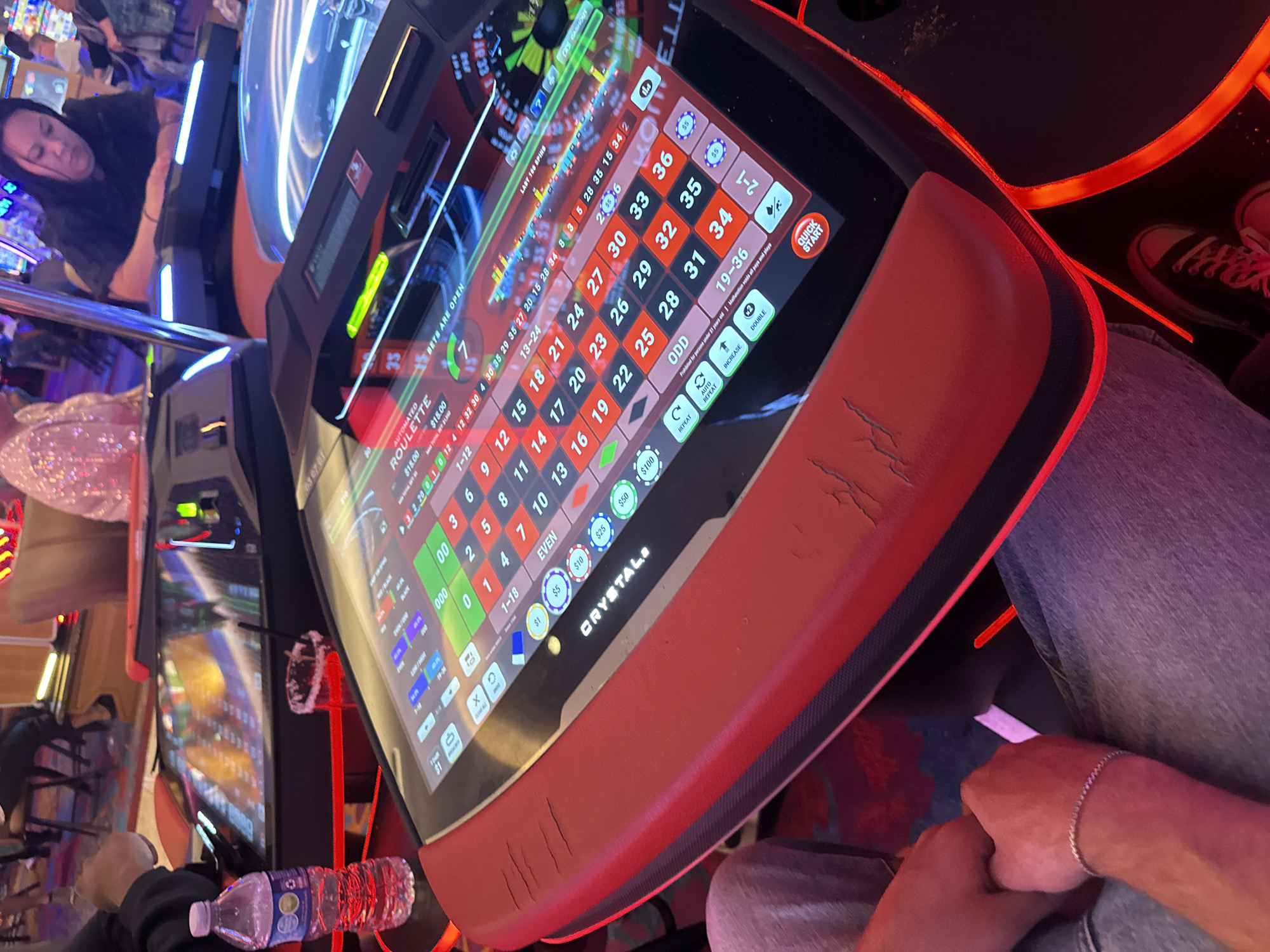 The image shows a person playing an automated roulette machine. The machine is branded "CRYSTAL". The screen displays the roulette layout with betting options and recent spin results. Casino lights are visible in the background along with other patrons. The scene appears to be inside a casino, given the roulette machine and gambling setting. Based on the information on the machine, the location is Hollywood Seminole Reservation, United States of America.