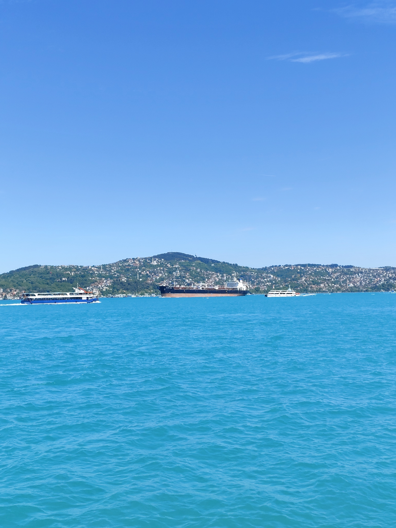 The image shows a wide expanse of water, likely the Bosphorus Strait, under a clear, bright blue sky. In the distance, there is a hilly landscape densely populated with buildings and trees, characteristic of the Sarıyer district in Istanbul, Turkey. A large tanker ship is positioned in the middle of the water, appearing to be stationary or moving slowly. Two smaller passenger ferries, one on the left and another on the right, are also present, moving across the water. The water is a consistent turquoise color, with gentle ripples reflecting the clear sky above.