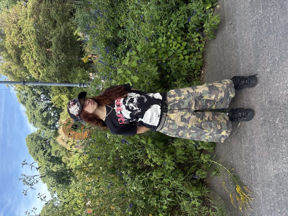 The scene depicts a young female standing and posing on a paved pathway within the Rosedal de Palermo park in Buenos Aires, Argentina. She has long, wavy, reddish-brown hair and is looking directly at the camera with pouting lips. She wears a dark baseball cap featuring light-colored graphics, a black short-sleeved t-shirt with a prominent white graphic of a stylized, grotesque figure and red/pink text, a silver-colored chain with a pendant, and a dark studded belt. Her lower attire consists of baggy, camouflage-patterned pants with yellow star motifs, paired with dark, chunky sneakers or boots. The background is dominated by dense green foliage, including shrubs with small blue/purple flowers and large trees. A wooden architectural element with an arched opening and latticework is visible among the trees in the mid-left. A dark, slender lamppost is present on the right side, behind the subject. Faint, blurry human figures are discernible in the far background. On the paved ground in the lower left, a cluster of yellow flowers lies. The setting is outdoors, under bright daylight.