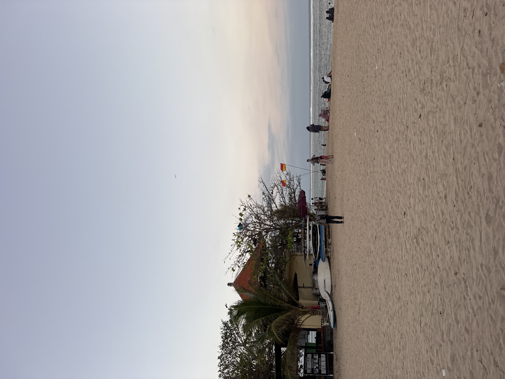 This image depicts a serene beach scene in Kuta Selatan, Indonesia, at dusk. The sand is golden and smooth, with subtle ripples indicating the receding tide. In the foreground, the beach stretches out towards the ocean, with a few scattered pieces of litter visible.
On the left side of the image, a small building with a red-tiled roof serves as a prominent landmark. In front of it, several surfboards and a small boat are neatly arranged on the sand, suggesting a surf school or rental shop. A palm tree stands tall beside the building, its fronds swaying gently. A sign with various offerings like "HAIR BRAIDING," "MASSAGE," and "TATTOOS TEMPORARY" is visible to the left of the building.
Further back, towards the water's edge, a group of people are scattered along the beach. Some are walking, while others are seated, enjoying the tranquil evening atmosphere. Two red and yellow flags are planted on poles, indicating the presence of a designated swimming area or safety warnings. The ocean is calm, with gentle waves rolling onto the shore. The sky is a soft gradient of pale blue and pink, transitioning into a dusky twilight, with a faint airplane visible in the upper left quadrant of the sky. The overall impression is one of a peaceful and relaxed beach environment.