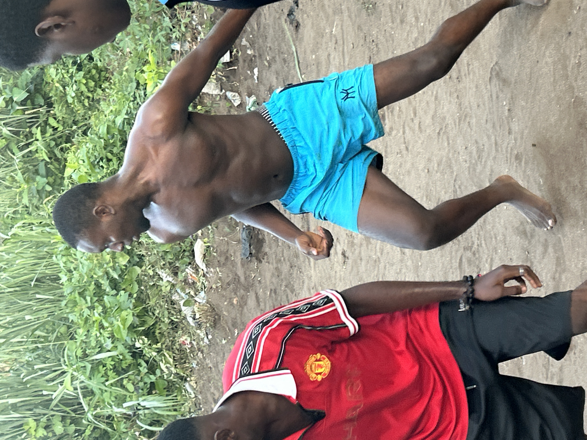 Three young Black men are in a sandy area with lush green vegetation in the background. One man, on the left, is wearing a red Manchester United jersey with white and black trim and black shorts. He has a beaded bracelet on his left wrist and a ring on his right index finger. The second man, in the center, is shirtless, wearing turquoise athletic shorts with a black and white striped waistband. He is looking to his left and appears to be in motion, with his arms slightly extended. The third man, on the right, is mostly obscured, but his face and shoulder are visible, and he appears to be looking at the center man. The environment suggests an outdoor setting, possibly near a body of water or a riverbank, given the sandy ground and dense vegetation. The scene is likely in Ihiagwa, Nigeria, a region known for its natural beauty and vibrant communities.