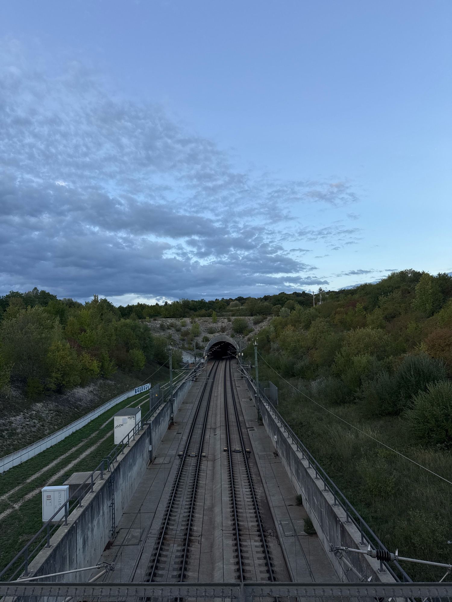 The image shows a high-angle view of a railway line leading towards a tunnel entrance in Wettstetten, Germany. The sky is a pale blue with scattered clouds. On either side of the tracks, there are lush green trees and vegetation. The railway is elevated on a concrete embankment, with railings along the sides. There are utility boxes and some graffiti on the embankment walls. The tunnel entrance is dark and appears to be illuminated from within. The scene is calm and devoid of any people or vehicles.