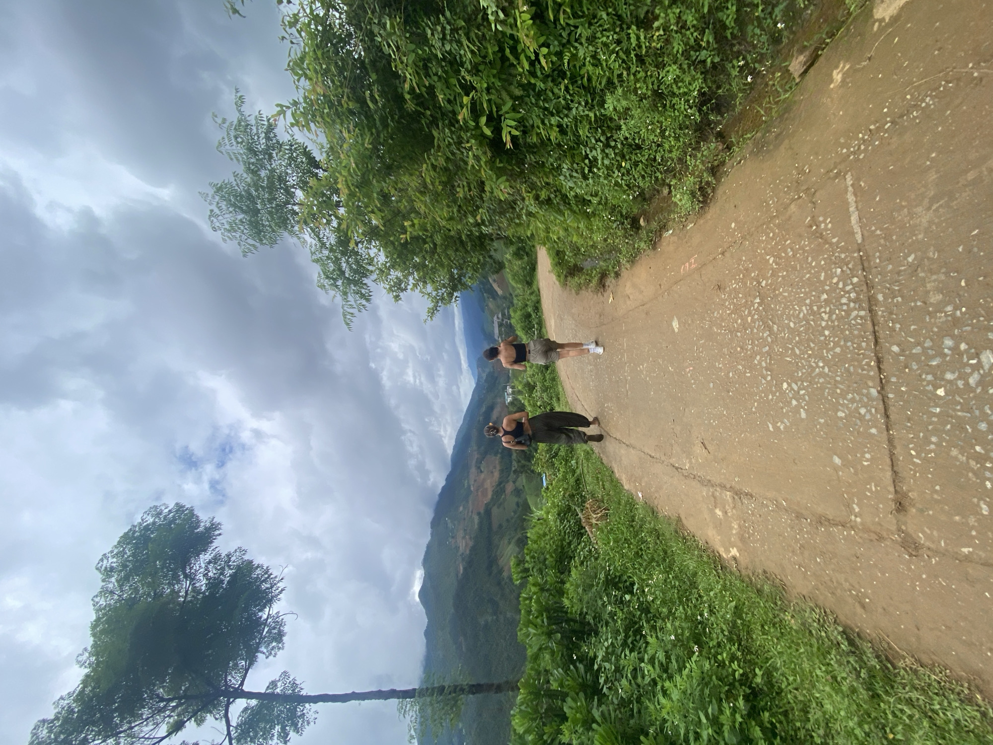 Two individuals, observed from behind, walk along a reddish-brown dirt and gravel path winding through a lush, mountainous landscape in Dế Xu Phình, Vietnam. The person on the left wears a dark sports bra or crop top, dark baggy pants, and dark footwear, carrying a dark shoulder bag. The person on the right wears a dark sports bra or crop top, light-colored shorts, and white sneakers with white socks. Both appear to be female and have dark hair.
The path, uneven with visible stones and cracks, is flanked by dense green vegetation. On the left, a tall, slender tree with a prominent canopy stands, while the ground is covered in abundant green bushes and smaller plants, including some small white flowers near the path's edge. The right side of the path features a thick wall of dense green foliage, comprising bushes and trees.
In the background, a series of green mountains with visible terraced or cultivated sections displaying patches of reddish-brown earth rise under an overcast sky filled with numerous grey and white clouds. The distant mountains are partially obscured by haze, suggesting a humid environment. The overall scene depicts a hike or walk through a vibrant natural setting.