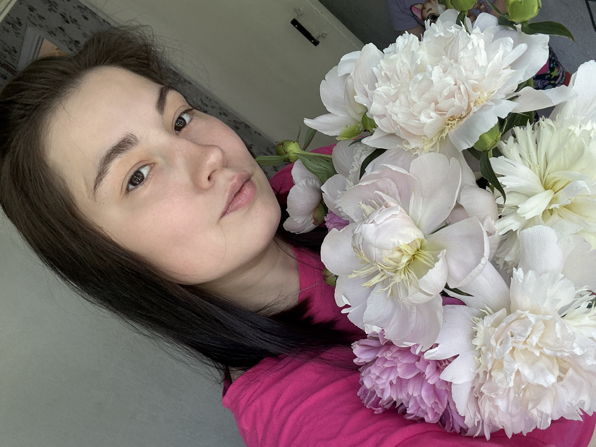 A young woman with dark hair and fair skin is holding a bouquet of peonies. She is wearing a bright pink t-shirt and a silver necklace. The peonies are a mix of white and pink, with some buds also visible. The background is a plain wall and a partially visible doorway. The image is taken from a close-up perspective, focusing on the woman's face and the flowers.