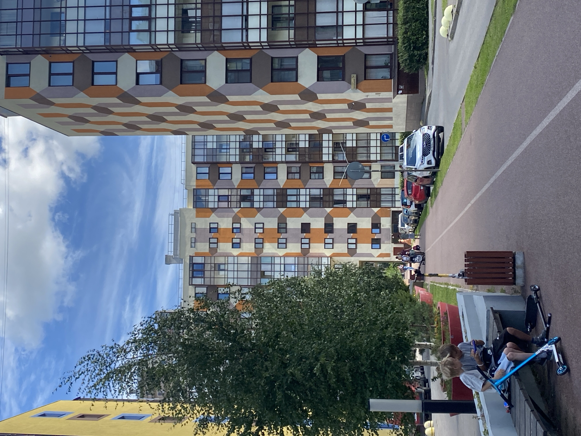 In Kudrovo, Russia, a modern residential courtyard is depicted under a bright, partly cloudy sky. The foreground features two young boys sitting on a wooden bench, engrossed in a portable game console. Scooters are present near the bench, suggesting outdoor play. A red paved pathway for cycling or walking runs parallel to the buildings, with a wooden waste bin nearby. The scene is framed by lush green trees and vibrant red planters.
Dominating the background are multi-story apartment buildings with distinctive geometric facade patterns in shades of orange, brown, and gray. The architecture is contemporary, characterized by large, reflective windows. Cars are parked along the street beside the buildings. The overall impression is one of a well-maintained, contemporary urban residential area designed for communal living and recreation.