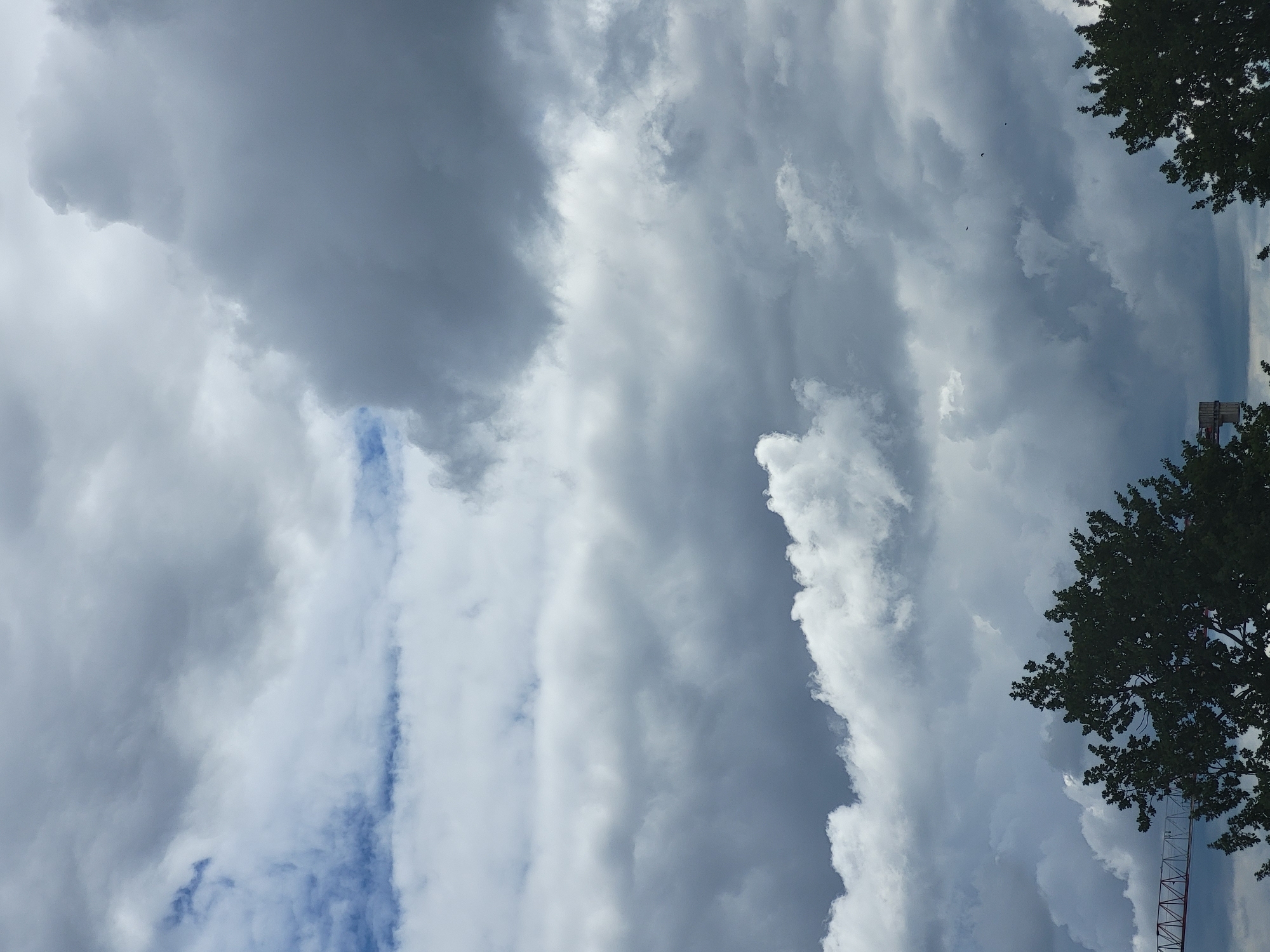 A low-angle shot captures a dramatic sky filled with thick, gray, and white clouds. A sliver of blue sky is visible near the top left. In the foreground, the dark green tops of trees are visible, and to the left, the red and white latticework of a construction crane extends into the frame from the bottom left edge. The overall scene suggests an overcast day with a hint of building structures.