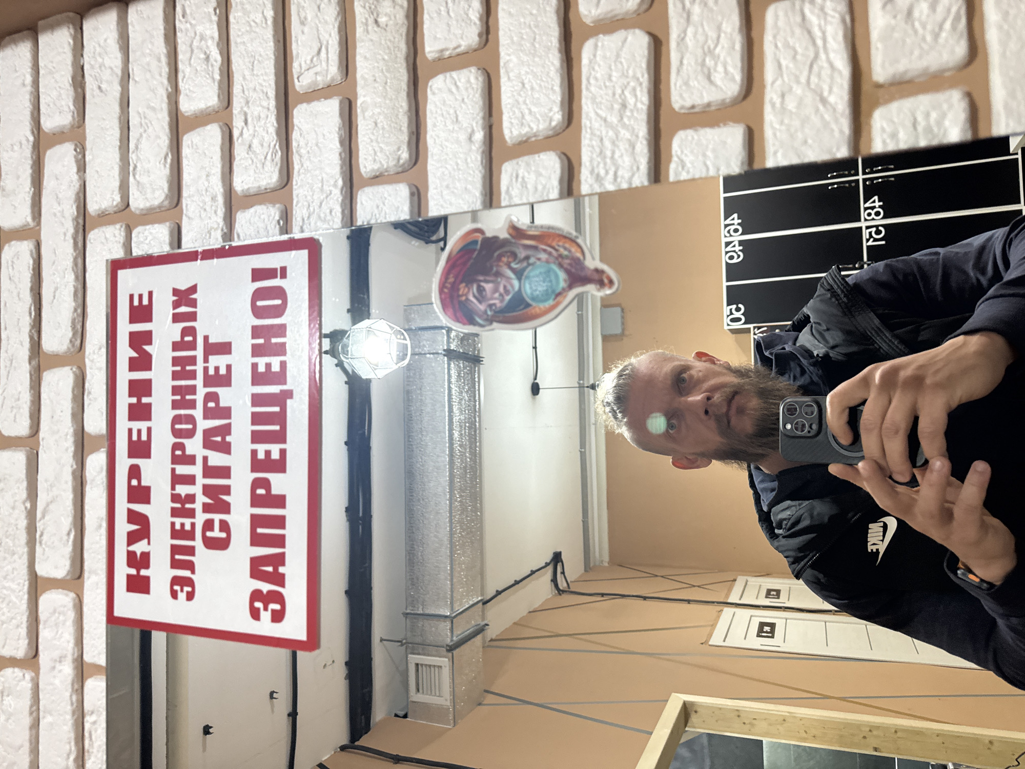 This media file captures a selfie taken by a man in a mirror, with the image rotated 90 degrees clockwise. The man, seen in reflection, has a beard and fair hair, and is wearing a dark jacket with a visible white "NIKE" logo on the sleeve. He is holding a dark smartphone, likely an iPhone model with multiple cameras, to capture his reflection.
The setting appears to be an indoor space in Volzhsky, Russia. Above the mirror, a wall is decorated with white, rectangular brick-like tiles on a light brown background. The mirror reflects various elements of the room. On the left side of the reflection, a prominent red and white sign is visible, displaying Cyrillic text that translates to "SMOKING ELECTRONIC CIGARETTES IS FORBIDDEN!"
Further reflections include a brightly lit, geometric light fixture near the center, and a colorful sticker of a stylized face, possibly a fortune teller, holding a glowing orb. To the right of the man's reflection, a black board with white numbers ("20," "40," "84," "21," "18") is visible. The general environment suggests an industrial or utility area, or perhaps a space undergoing renovation, with visible metallic ductwork, various electrical wires running along walls and the light brown floor, and white doors reflected in the distance. The lighting is artificial.