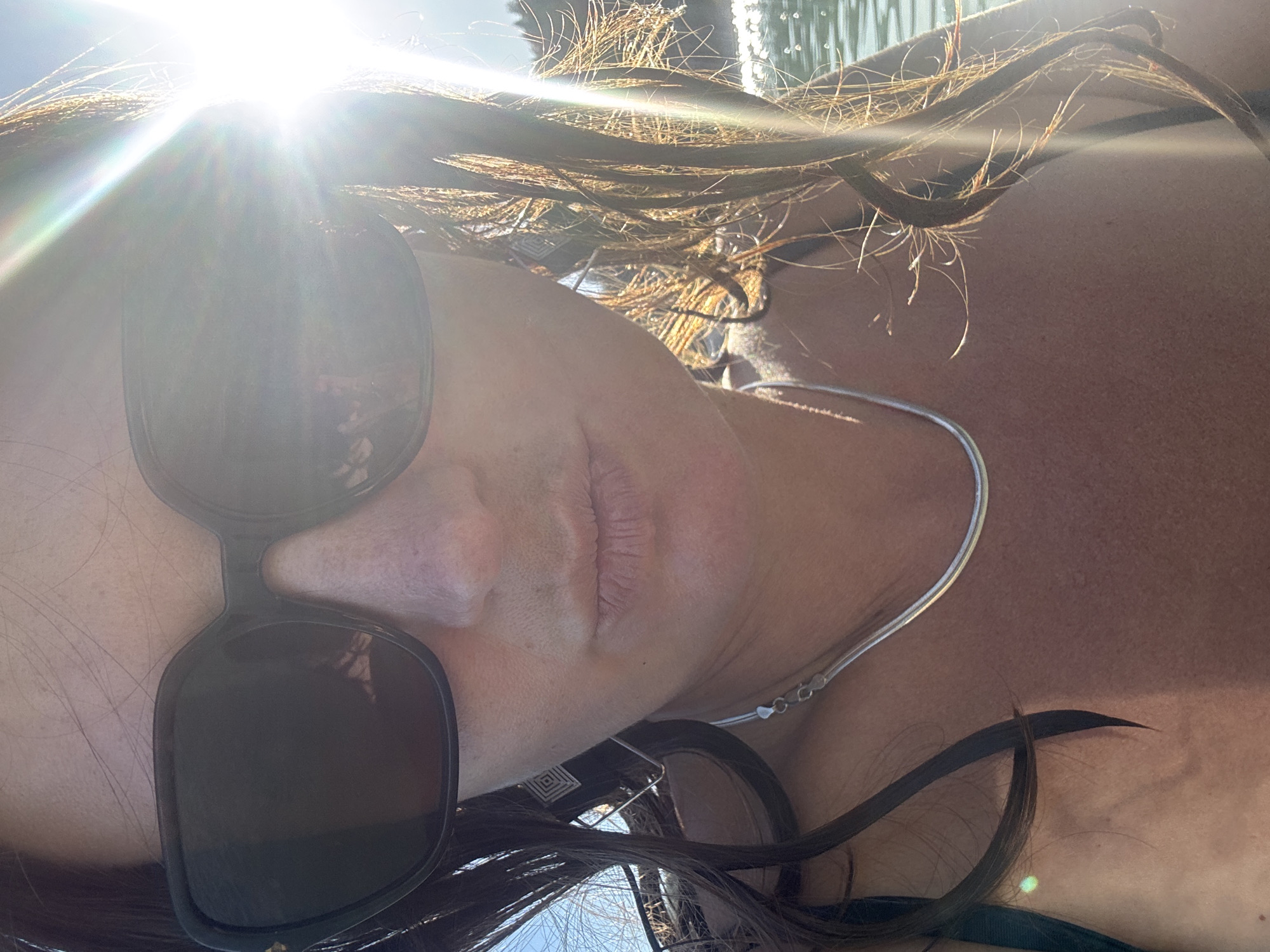 A close-up selfie of a woman wearing large, dark sunglasses, obscuring her eyes. She has dark hair that frames her face and extends over her shoulders. Her lips are slightly parted, giving the impression she is about to speak or has just finished. A delicate silver necklace is visible around her neck. The background is blurred and appears to be a body of water with sunlight glinting off the surface, suggesting an outdoor, possibly coastal or lakeside, setting. The sun is bright and positioned to the right of the frame, causing lens flare and highlighting strands of her hair. There is no discernible indication of the location being Maisod, France.