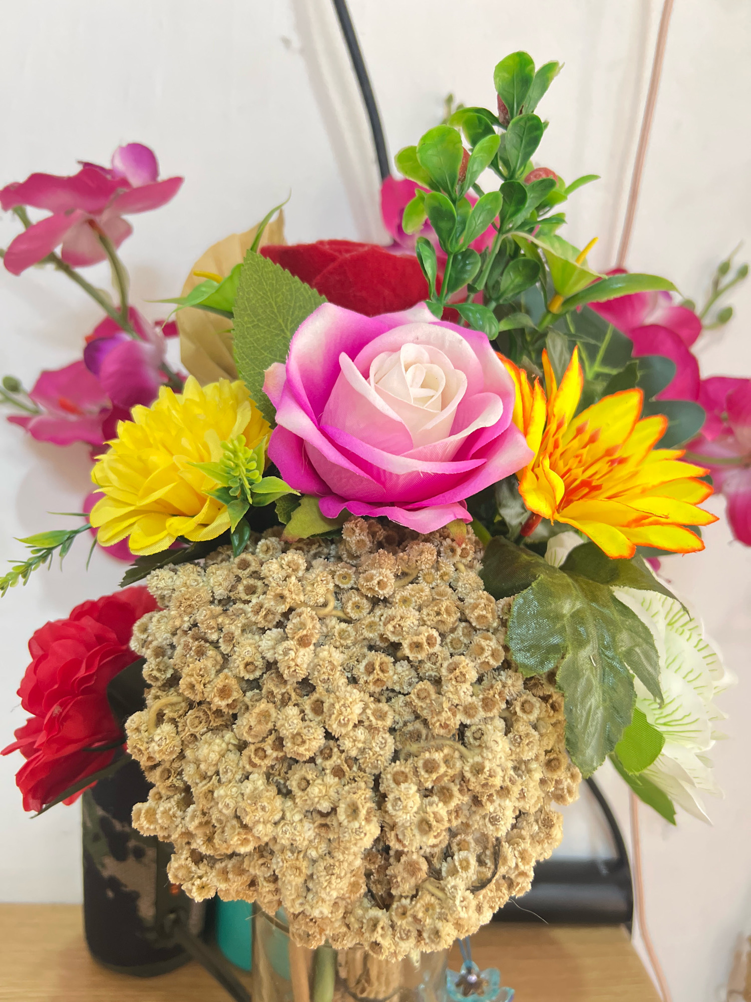 A close-up, vertical shot displays a floral arrangement in a vase. The arrangement is comprised of artificial flowers, including a large pink rose with white accents, a bright yellow chrysanthemum, a red rose, and yellow flowers with red streaks. There are also clusters of small, dried, tan flowers and green foliage. The background is a white wall. The bottom of the image shows a wooden surface, suggesting the arrangement is on a table or shelf. The lighting is bright, with some shadows cast on the wall. No people are present in the image.
