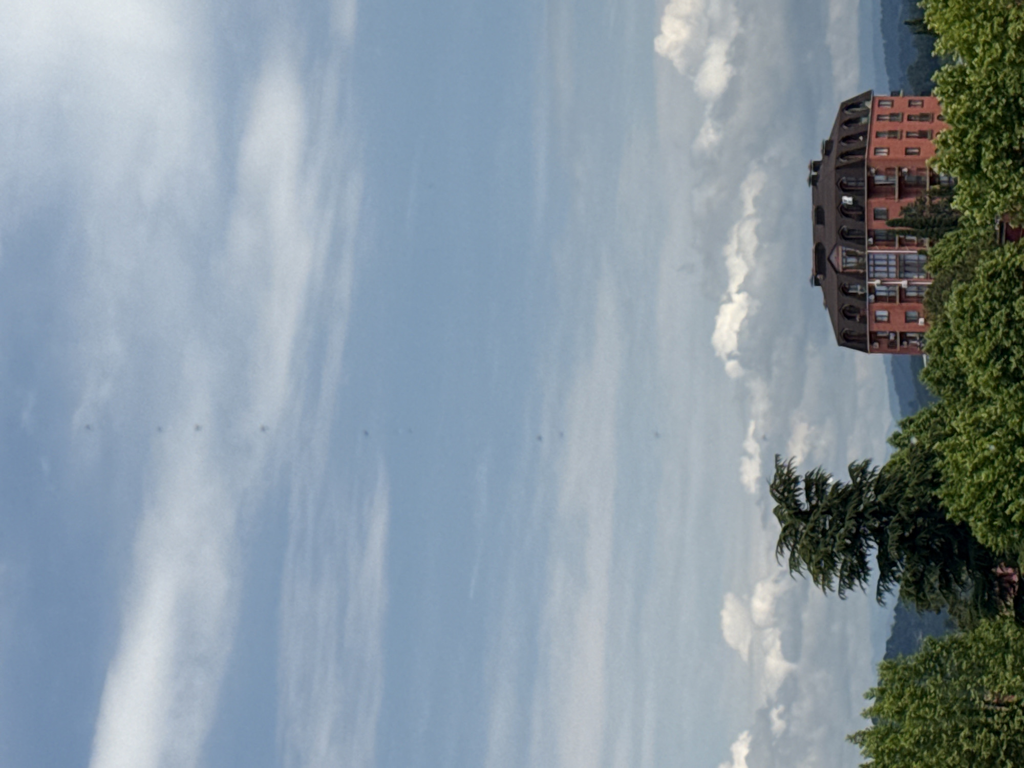 This is a medium shot of a residential building in Sochi, Russia, with a clear sky and scattered clouds. The building is a multi-story structure with a distinctive mansard roof. It is red-brick with multiple windows and balconies. In the foreground, there are lush green trees, including a tall, mature conifer tree. The background shows rolling hills, suggesting a natural landscape surrounding the city. The image appears to be taken from an elevated position, offering a panoramic view.