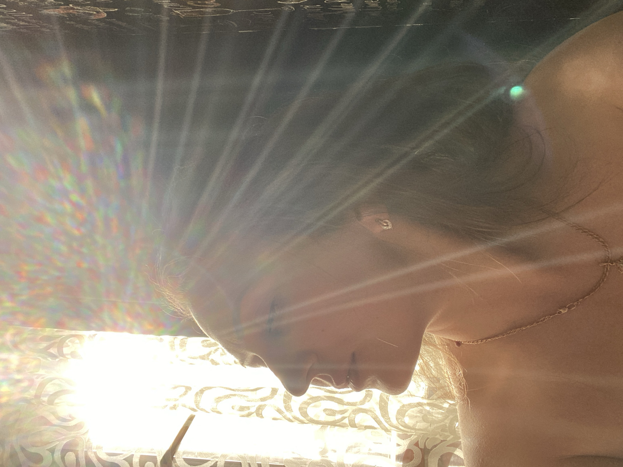 The image shows a woman's face in profile, illuminated by sunlight shining through a patterned curtain. The light creates a rainbow effect. The woman has fair skin, dark hair, and is wearing a necklace. An earring is also visible. The background is dark and appears to be a ceiling or wall. There is no information available to determine the location in the city of Марковский, Russia.