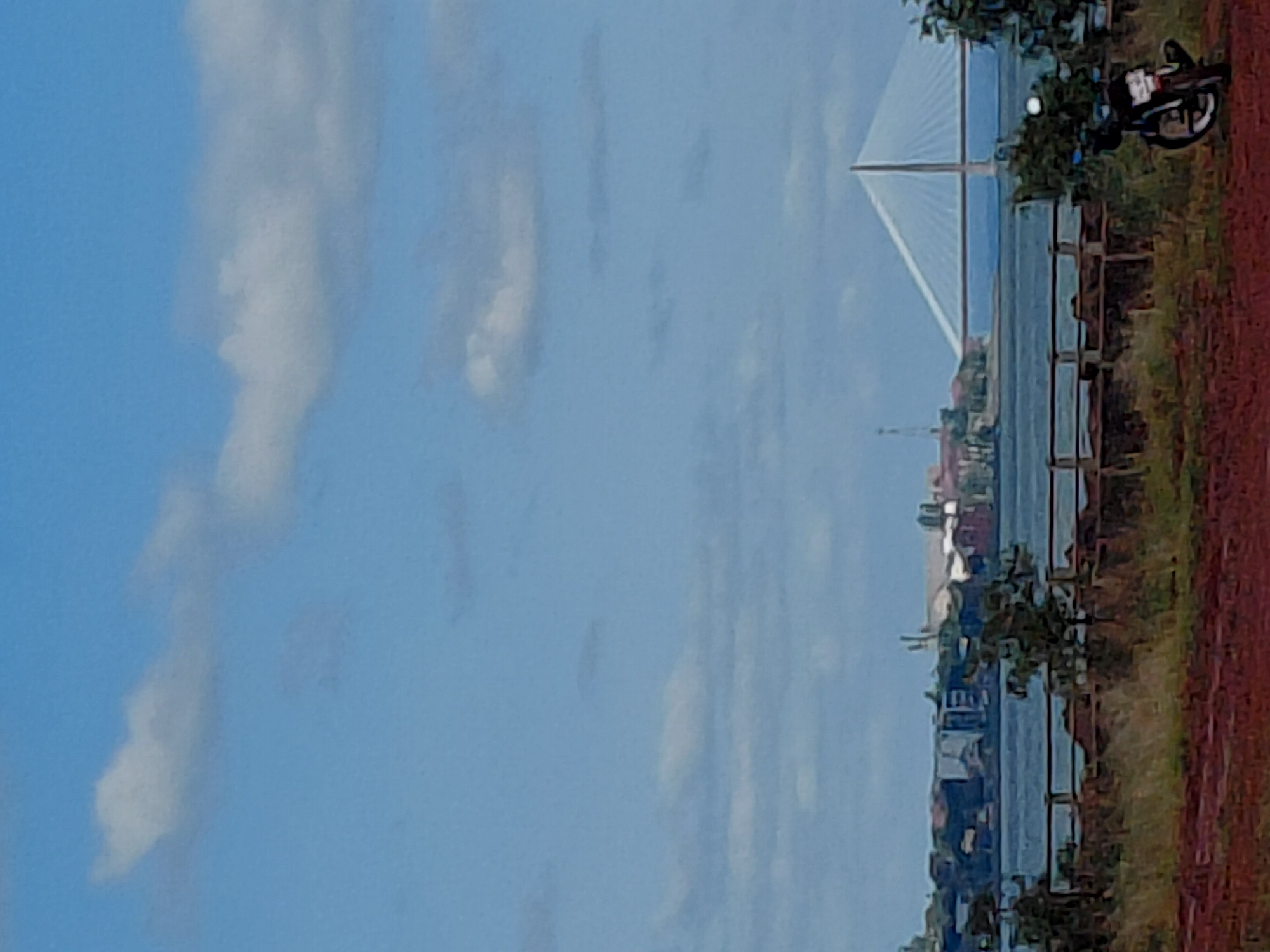 The image shows a scenic view of the city of Encarnacion, Paraguay, across the Parana River. A large suspension bridge, likely the Posadas-Encarnacion Bridge, dominates the mid-ground. The bridge's white cables fan out from a central pylon, creating an impressive architectural feature. On the opposite bank, a cluster of buildings, possibly industrial or port facilities, are visible with some smokestacks. The foreground features a grassy embankment with a wooden fence, and a motorcycle is parked on the right side of the frame. The sky is a bright blue with scattered white clouds. The overall scene suggests a sunny day with calm weather.
