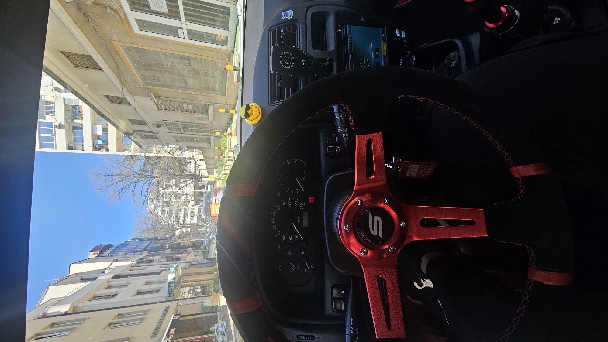 A view from inside a vehicle shows a car's dashboard and steering wheel, with a cityscape visible through the windshield. The steering wheel is black with a red rim and has a stylized, red "S" logo in the center. Various car instruments and controls are visible on the dashboard. Through the windshield, there is a view of a city street, with buildings and trees lining the road. This is Varna, Bulgaria.