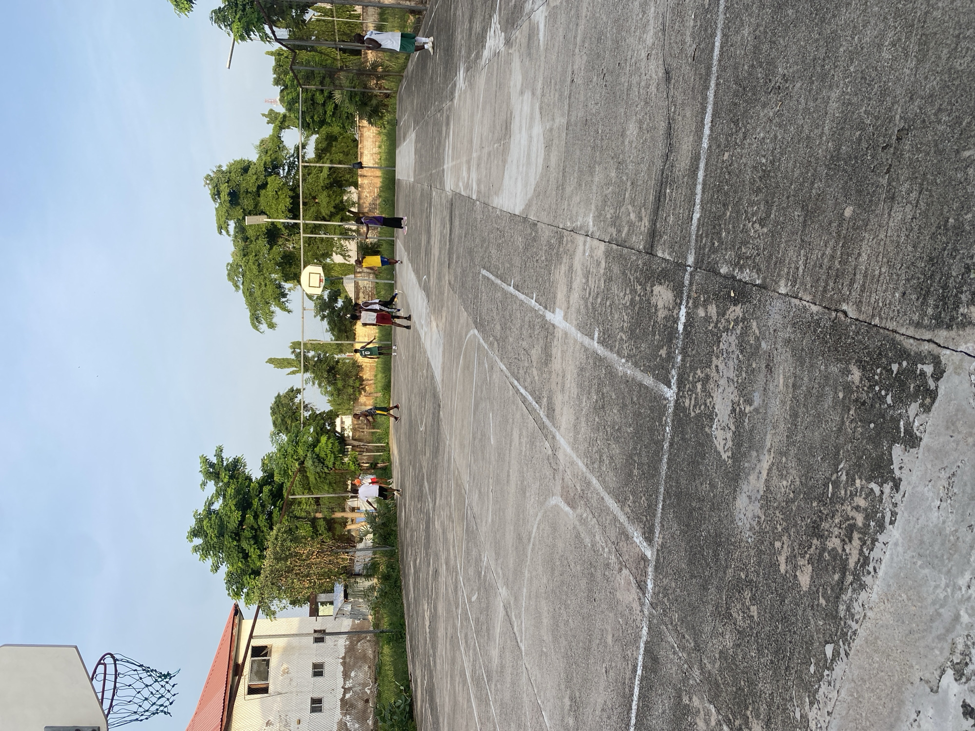 A group of young people are playing basketball on an outdoor court in Jalingo, Nigeria. The court is made of concrete and has white lines marking the boundaries and key areas. The sun is low in the sky, suggesting it is either early morning or late afternoon, casting long shadows across the court. The weather appears to be clear and warm, typical for the region.
Several individuals, appearing to be teenagers or young adults, are actively engaged in the game. Some are in motion, dribbling or running, while others are standing, observing or waiting for their turn. They are dressed in casual athletic wear, with some wearing jerseys of different colors. The scene evokes a sense of friendly competition and recreation.
In the background, lush green trees line the perimeter of the court, indicating a verdant environment. Beyond the trees, there are some buildings, one of which is a white structure with a red roof. A chain-link fence surrounds the court, with a basketball hoop and net visible on one side, along with a portion of a backboard. The overall atmosphere is one of casual outdoor activity in a community setting. No visible text is present in the image.