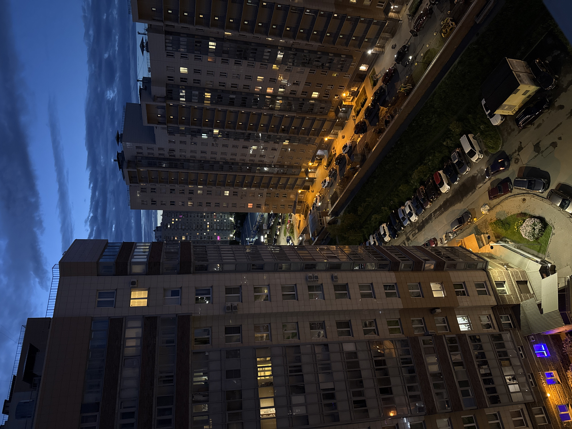 An aerial shot captures a residential city area in the late evening twilight. Dominating the foreground are the multi-story apartment buildings on the left and right sides of the frame, casting long shadows. Several windows in the buildings are illuminated, indicating occupancy. A street runs between the buildings, with parked cars lining its sides. The sky above is a deep blue, transitioning from lighter hues near the horizon to darker shades, with scattered clouds adding texture. The overall impression is one of urban development at dusk.