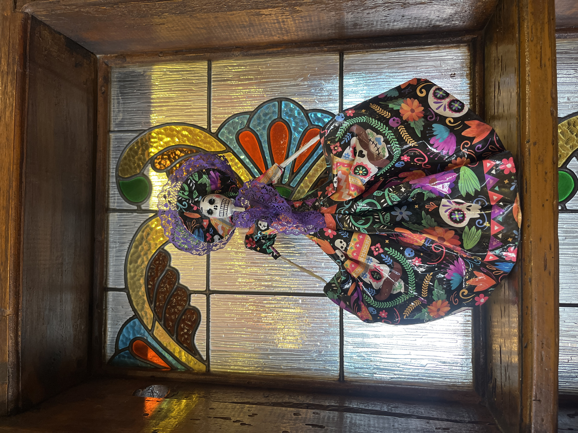 A Day of the Dead Catrina doll is propped against a leaded glass window with colored panes in a wooden frame, which appears to be located in Puebla, Mexico. The doll has a skull face and wears a black dress decorated with colorful skulls, flowers, and guitars. A purple lace veil adorns its head. The glass window consists of rectangular panes, and the leaded design incorporates yellow, green, brown, blue, and orange sections. The wooden frame is aged and shows signs of wear. The surface below the window is glossy and reflects light.