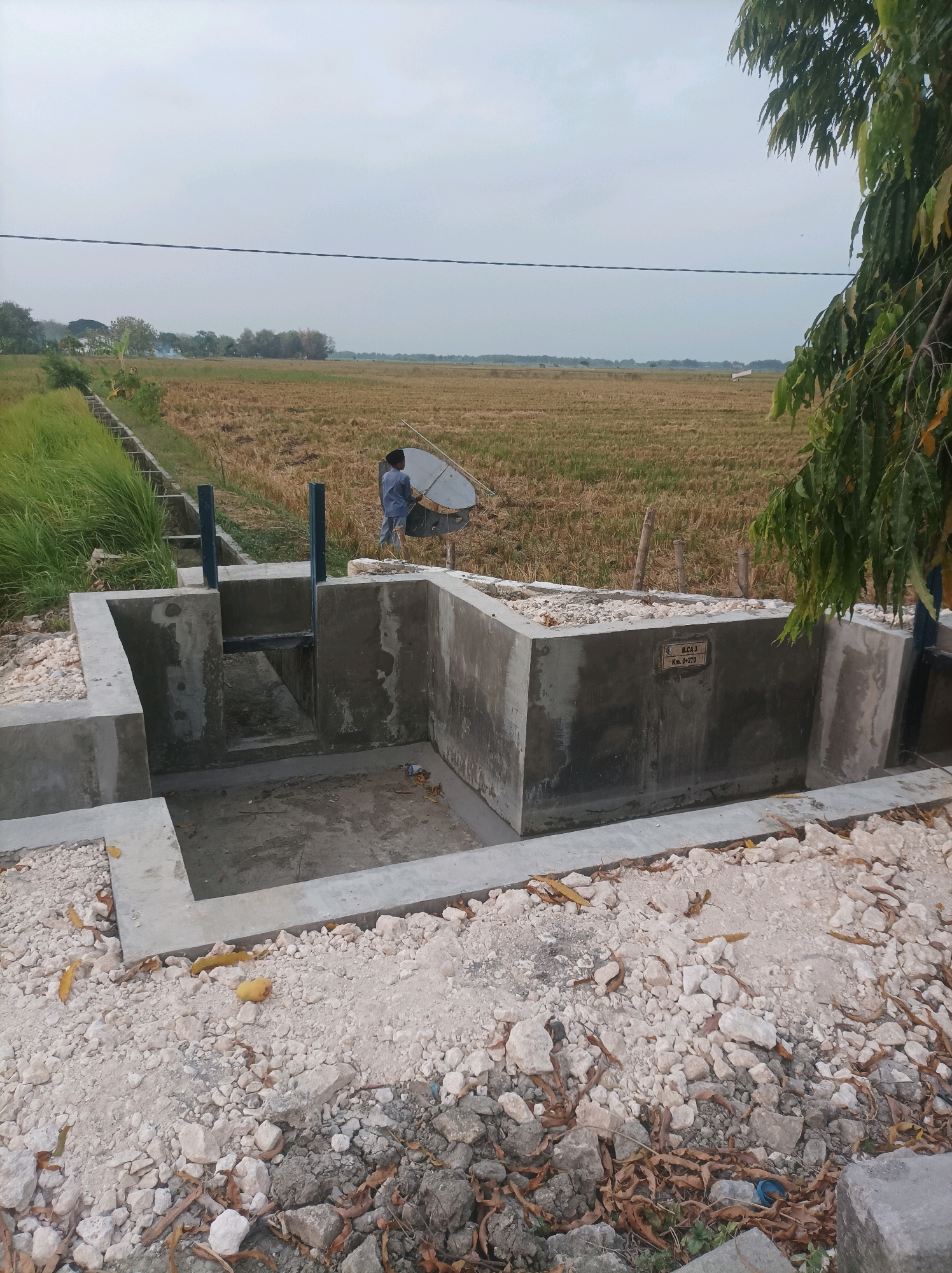 In CANGA'AN, Indonesia, an overcast sky looms over a rural landscape. In the foreground, a newly constructed concrete structure, possibly a water management system, is visible. It features several chambers and metal sluice gates. The ground surrounding the structure is covered with a mix of concrete rubble, small white stones, and dry leaves. A person dressed in a blue shirt and dark pants is standing near the concrete structure, holding a large, circular, metallic object, possibly a reflector or a part of a tool. Beyond the structure, a field of harvested rice stalks stretches towards the horizon, with a narrow irrigation channel running along the left side of the image. The environment suggests a setting outside of a typical urban center, with agricultural land dominating the background. A metal plaque is affixed to one of the concrete walls, bearing the inscription "B.C.A. 3 Km. 0+270".