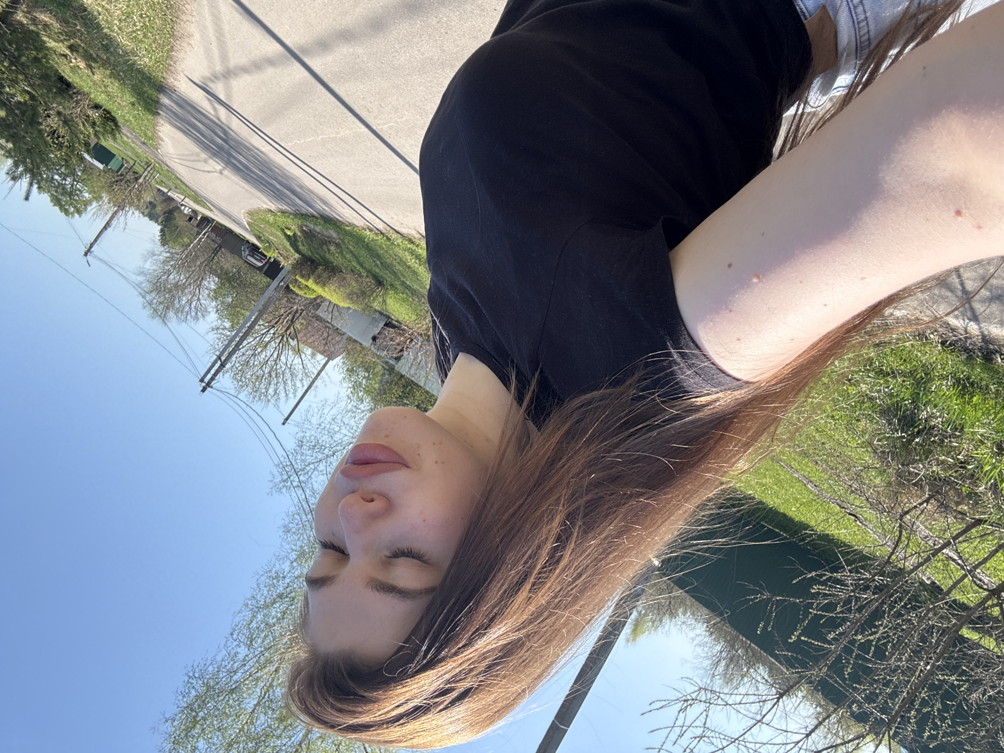 A woman with light skin and long brown hair is centered in the foreground, wearing a black shirt and denim jacket. Her eyes are closed. The background includes a street, grass, trees, and a blue sky. There is no evidence to suggest the location is in the city of Мещёры, Russia.