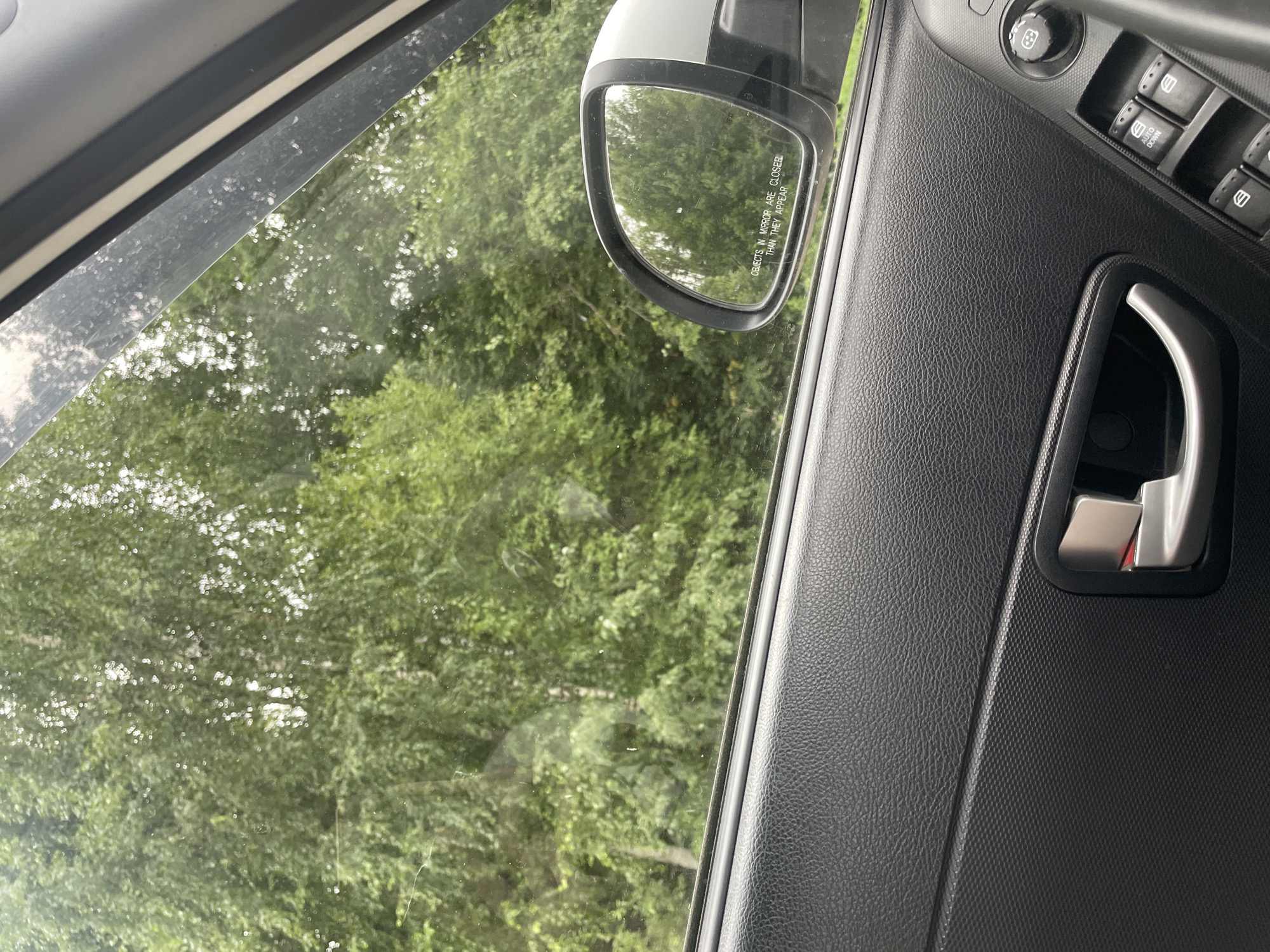 An indoor shot captures a car's interior from the passenger side, looking out through the window. Lush green trees and foliage fill the exterior view. A side mirror is visible, reflecting the same green scenery and displaying the warning "OBJECTS IN MIRROR ARE CLOSER THAN THEY APPEAR." The car's black textured door panel, interior door handle, and a window control panel with buttons are prominent in the foreground. The scene is set in Gavrilovka, Russia.