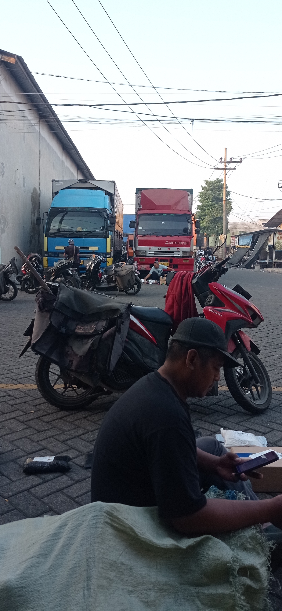 In the city of Jemundo, Indonesia, a roadside scene unfolds with several vehicles and individuals. The foreground features a man wearing a black t-shirt and a dark baseball cap, sitting on the ground, looking at his smartphone. He is positioned next to a dark-colored motorcycle with a large bag attached to its rear. To his right, a red Honda motorcycle is parked, with a red cloth draped over its seat.
Behind the motorcycles, a red Mitsubishi Fuso truck is visible. A person in a light blue shirt is interacting with items on the ground in front of the truck. Further back, a blue truck with a yellow stripe is parked, with a person standing near its rear. The ground is paved with interlocking bricks. In the background, utility poles and wires crisscross the sky, suggesting an urban or semi-urban setting. The overall atmosphere appears to be a busy loading or waiting area for transport vehicles.