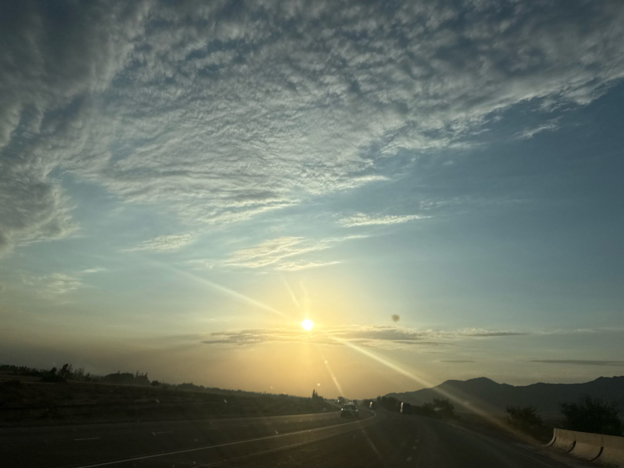 The image depicts a sunset scene viewed from a vehicle traveling on a highway. The sun is setting in the distance, casting a warm, golden light across the sky and illuminating the clouds with streaks of yellow and orange. The sky is a gradient of blue and yellow, with scattered clouds creating interesting patterns. The landscape appears dry and arid, with low-lying hills or mountains in the background and sparse vegetation. Several vehicles are visible on the highway, suggesting ongoing traffic. The location is identified as the city of Chüy, Kyrgyzstan, implying this highway is in or near that region.