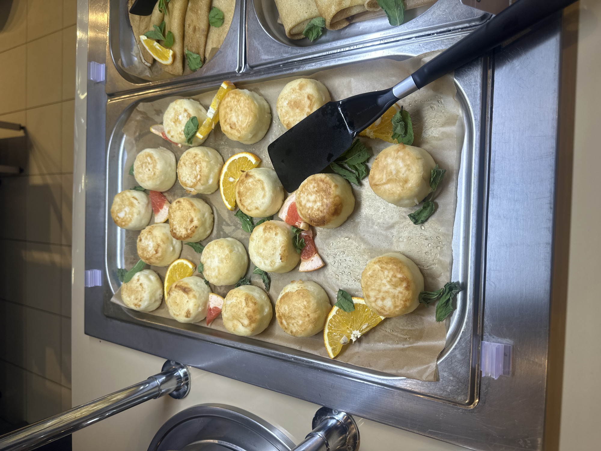 The image depicts a stainless-steel serving tray containing approximately twenty small, round pastries, likely Syrniki or similar cheese pancakes. The pastries are arranged in rows on a sheet of parchment paper. Orange and grapefruit segments, along with fresh mint leaves, garnish the pastries. A black spatula rests in the upper center of the tray. The tray is placed within a refrigerated display case. The location appears to be a buffet or food service area. It is not possible to confirm the city of Krasnaya Polyana, Russia, without additional information.