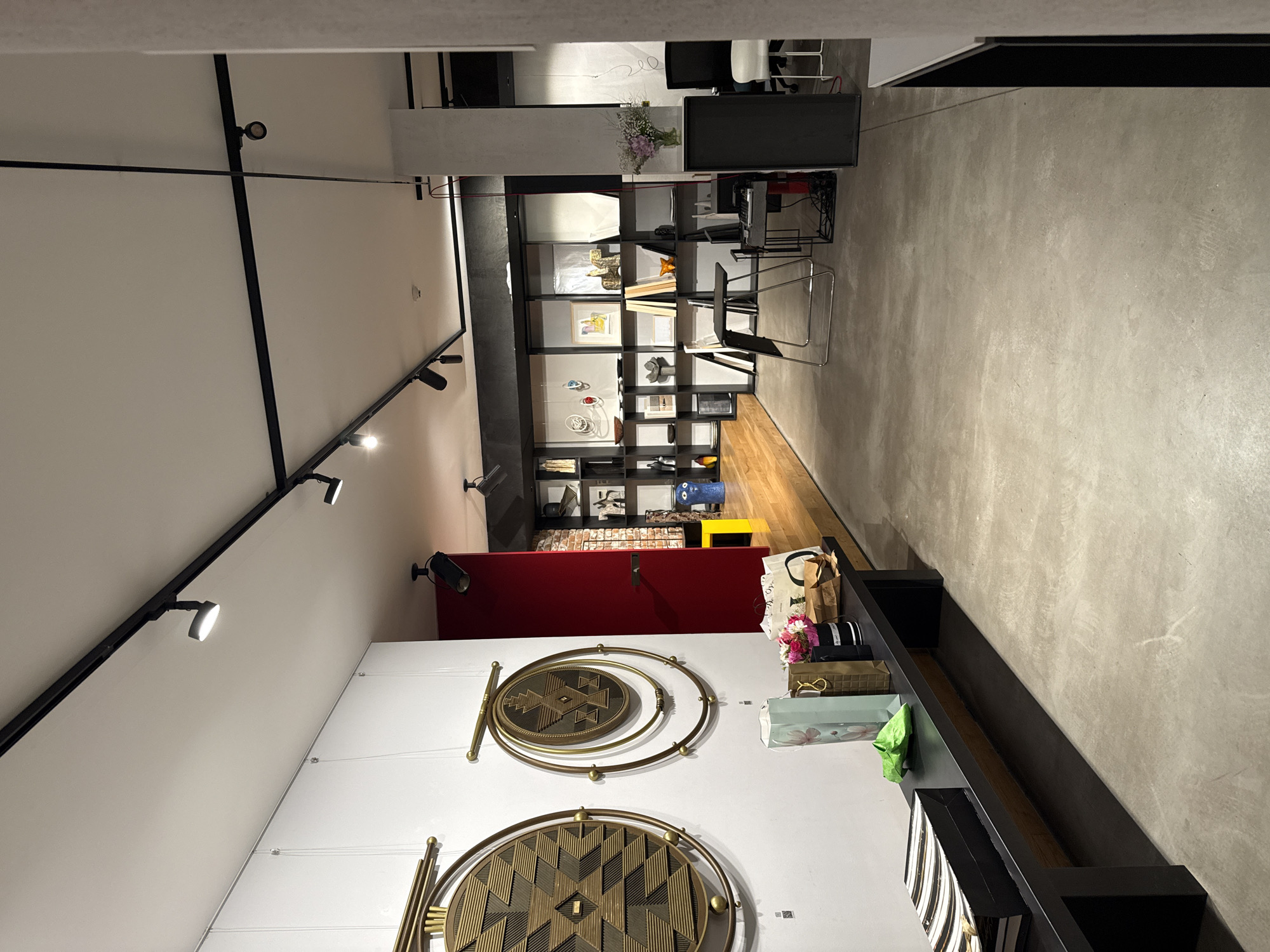 The media file captures a modern and meticulously arranged interior space, likely a showroom, art gallery, or contemporary office in Sofia, Bulgaria. The image is oriented vertically but depicts a horizontal layout of the room.
The space features a long, open plan with distinct material zones. On the right side of the image, a white wall prominently displays two large, circular brass-colored wall hangings, each intricately designed with geometric patterns. A long, dark-colored counter or display shelf runs along this wall, holding several paper bags, a small bouquet of mixed flowers, and decorative boxes, one with a small green cloth.
To the left, a track lighting system with multiple spotlights illuminates the white ceiling and walls, contributing to the bright and well-lit atmosphere. Further into the room, a section of the wall is painted a striking red. Below this, extensive black shelving units are filled with various decorative objects, sculptures, framed pictures, and books, suggesting an exhibition or retail display. A metallic, modernist-style chair (reminiscent of a Wassily chair) is positioned near these shelves.
In the background, a desk area is visible with a black computer monitor, a black office chair, and another white chair. The flooring transitions from a smooth, light gray concrete or polished surface on the left side to warm wooden planks deeper within the space. A vase of purple and white flowers is placed on a counter or partition near the desk area.
No people are present, indicating a static scene. The overall impression is one of sophisticated design and curated presentation, emphasizing aesthetics and modern functionality. There is no discernible text visible in the image.