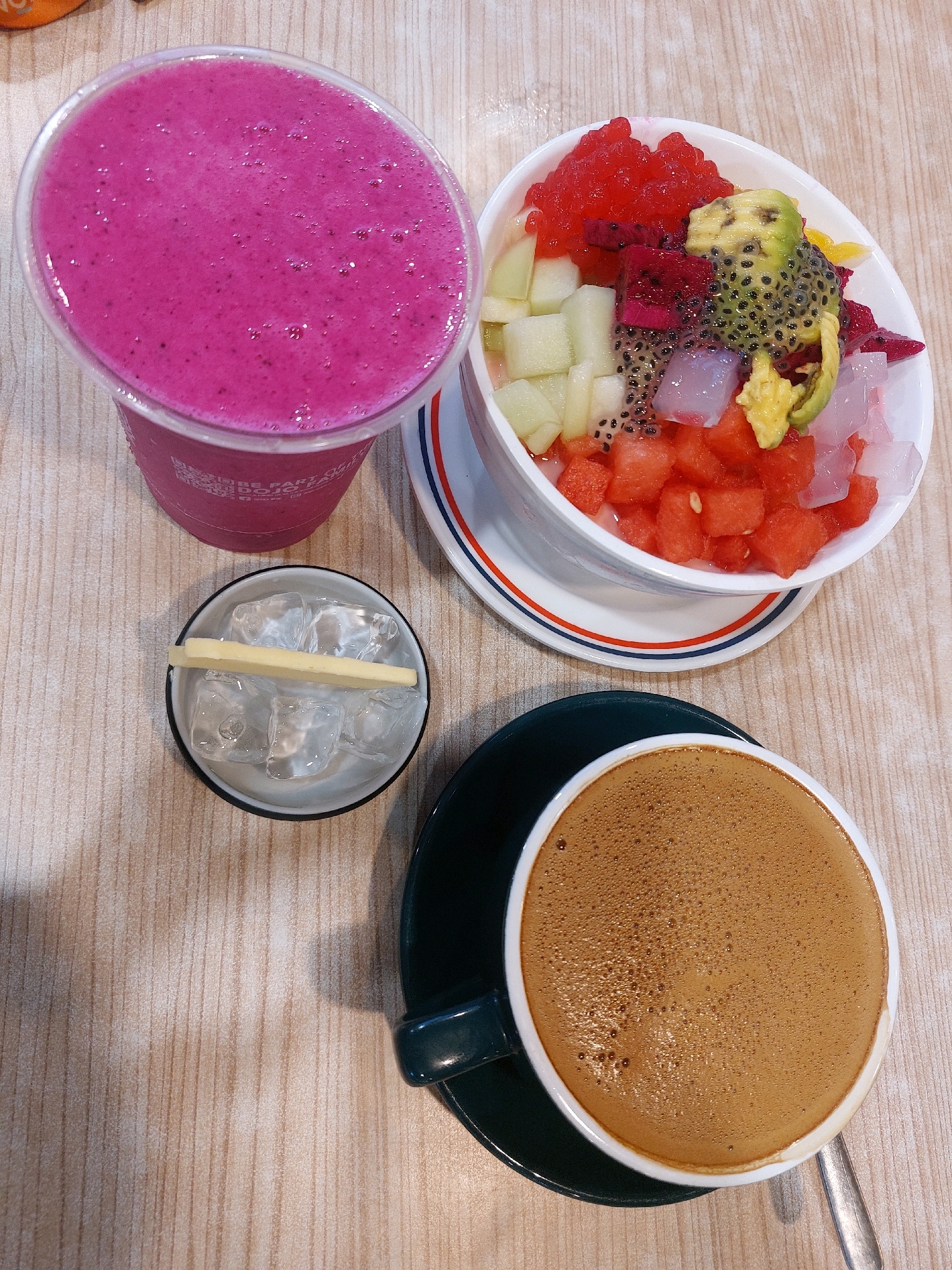 A top-down view of a table at a cafe in Tarakan, Indonesia, showcases a selection of beverages and a dessert. A tall, clear plastic cup filled with a vibrant pink dragon fruit smoothie occupies the upper left. To its right, a white bowl rests on a saucer with blue and red stripes, containing a colorful fruit salad. The salad is a medley of diced watermelon, cubes of light green melon, chunks of pink dragon fruit, and green avocado slices topped with black chia seeds and translucent cubes of jelly. A small, white dish containing ice cubes and a thin, yellow rectangular food item, possibly cheese or butter, is positioned below the smoothie. In the lower right corner, a white cup filled with a frothy, brown coffee sits on a dark green saucer, with a silver spoon resting to its right. The table's surface is a light-colored wood grain laminate.