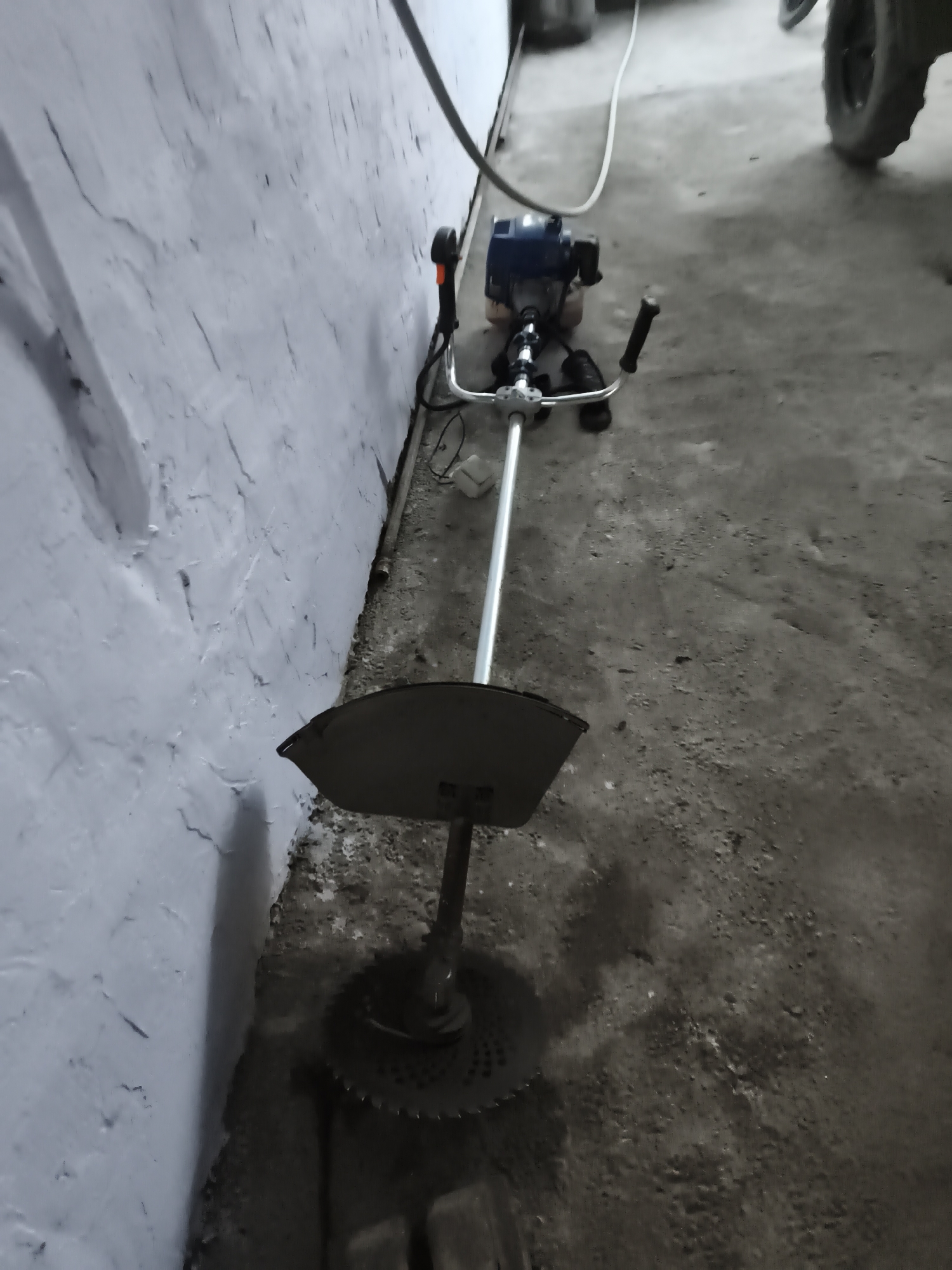 A gas-powered string trimmer with a blade attachment is positioned on a concrete floor next to a textured light-colored wall. The trimmer has a blue engine and a silver shaft with black handlebars. A dark circular guard and a metal blade are visible at the bottom. The wall has a rough, uneven surface with visible cracks and some dark smudges. The floor is concrete and appears slightly dusty. In the background, a white electrical cord runs along the floor. Part of a tire and a dark object are visible in the far right background. The lighting suggests an indoor environment, possibly a garage or storage area, in Beynəlmiləl, Azerbaijan. There are no people or animals present in the image.