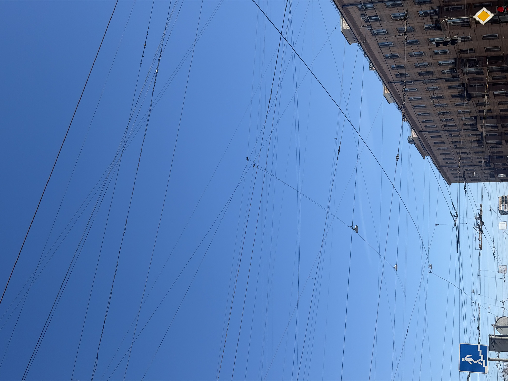 The image captures a clear, blue sky in Saint Petersburg, Russia. Power lines crisscross the sky, running from the side of a building on the right side of the image, where an emergency sign and a traffic light are visible.