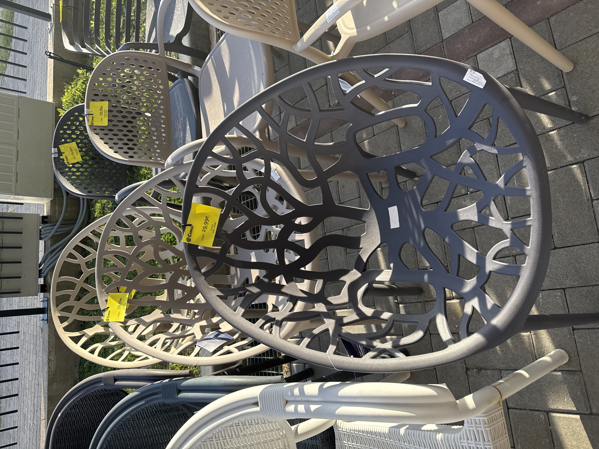 The image shows a collection of patio chairs displayed for sale. The chairs are made of plastic or a similar material, and they feature a variety of designs, including some with open-weave patterns and others with perforated seats. Price tags are attached to some of the chairs, indicating a price of 29.99. The chairs are arranged on a paved surface, possibly in a store or outdoor market in the city of Unknown, Unknown.