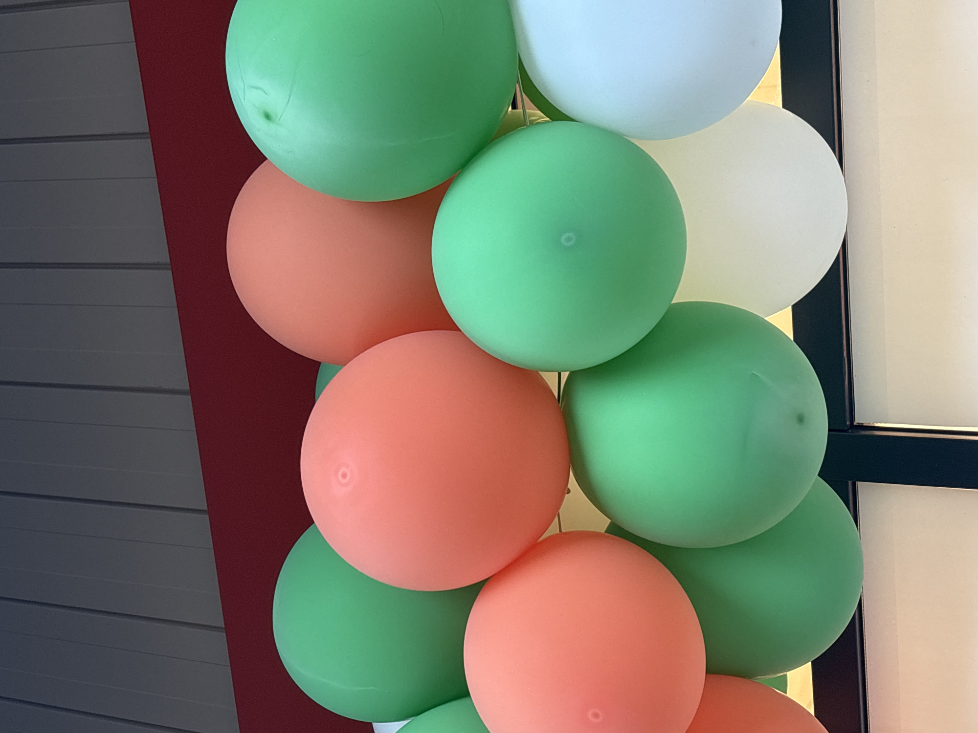 The image shows a close-up of a collection of inflated balloons of various colors, predominantly orange, green, and white. The balloons are clustered together, obscuring the background which appears to be a ceiling or upper wall structure with red and gray elements. The arrangement suggests a decorative display, possibly for an event. There are no people, animals, or clear indications of a specific city or location like Bila Tserkva, Ukraine within the image itself.