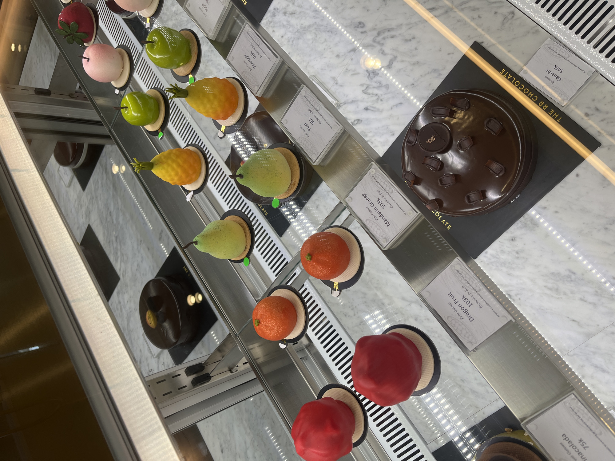 The image captures a brightly lit dessert display case inside a patisserie or chocolate shop, identified by the text "THE RR CHOCOLATE," likely situated in North Kuta, Indonesia. No people are visible in the scene.
The display features an array of meticulously crafted, fruit-shaped petit gateaux and a large chocolate cake, arranged on multiple shelves within a clean, modern glass and metal showcase. The individual petit gateaux, presented on small black and beige bases, include realistic representations of red berry, pink apple, green apple, green pear, yellow pineapple, reddish-orange mandarin orange, and red dragon fruit, each with a glossy finish.
On the right side of the display, a large, dark round chocolate cake, labeled as an "Entremet Ganache," is prominently displayed on a black base, decorated with small chocolate pieces and a circular branded element. Another smaller dark chocolate dessert is partially visible further down on the left.
The setting is indoors, with bright artificial lighting illuminating the desserts and creating reflections on the glass. The background of the display case appears to be white marble. The time of day and weather are not discernible from the indoor shot. The overall presentation aims to evoke appeal and desire for the confectionery.
Visible text on price tags includes: "THE RR CHOCOLATE" for the shop name. Dessert names and prices are "Entremet Ganache 545k," "Petit Gateau Dragon Fruit 103k," "Petit Gateau Mandarin Orange 103k," "Petit Gateau Pear 85k," "Petit Gateau Pineapple 103k," "Apple 75k," and "Pina Colada 75k." Many tags explicitly state "Exclusive in Bali," confirming the Indonesian location. A standard disclaimer reads, "All price are subject to prevailing government tax and service." The prices, using "k" for thousands, indicate Indonesian Rupiah.