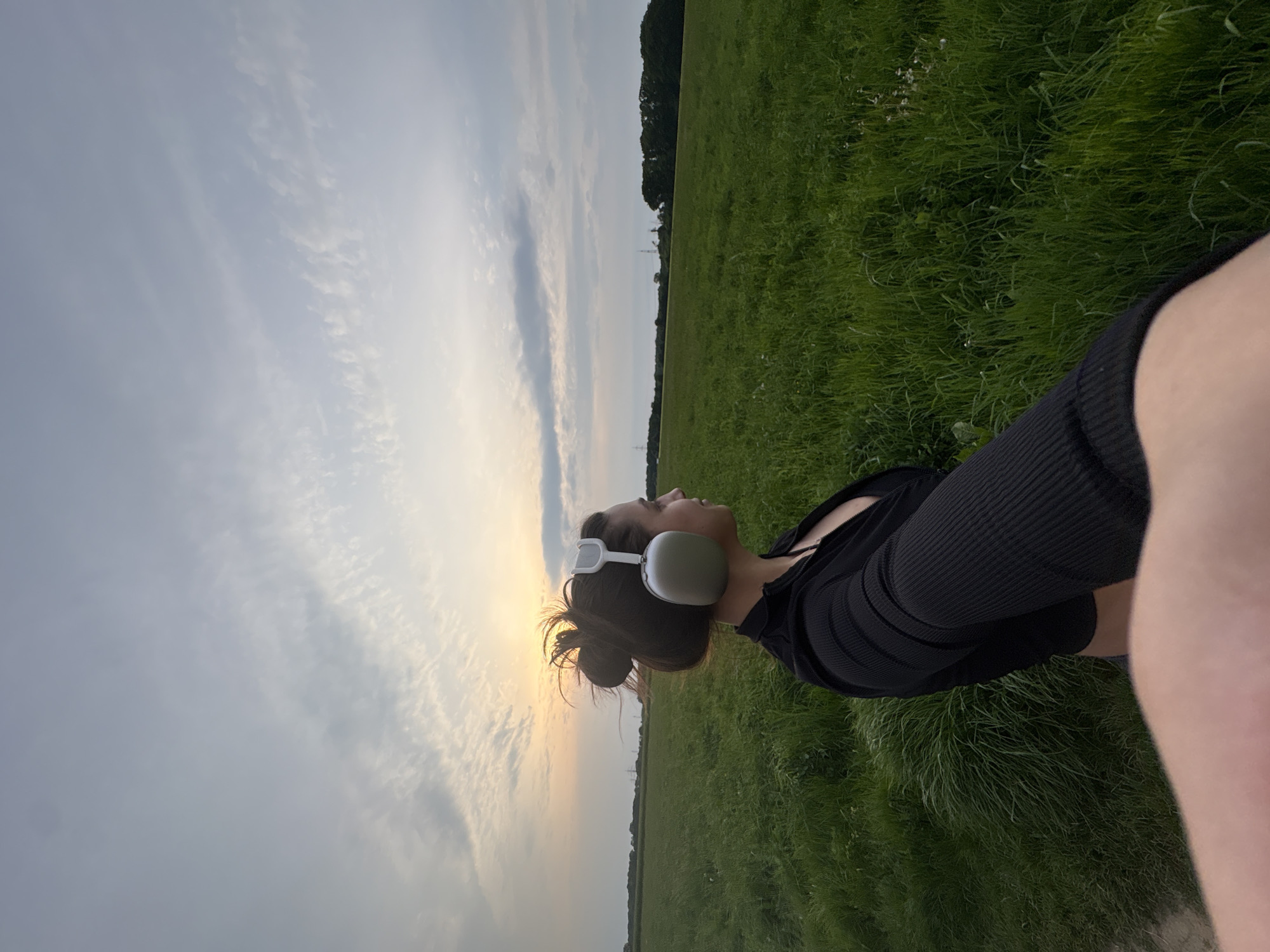 A woman is lying in a grassy field on what appears to be a slightly overcast day. The location is likely a rural area in Mönchengladbach, Germany. She is wearing a dark-colored, ribbed top. Headphones are over her ears. Her hair is gathered in a bun. Her face is turned towards the sky. The background includes a field of green grass. The sky is a mix of clouds and light.