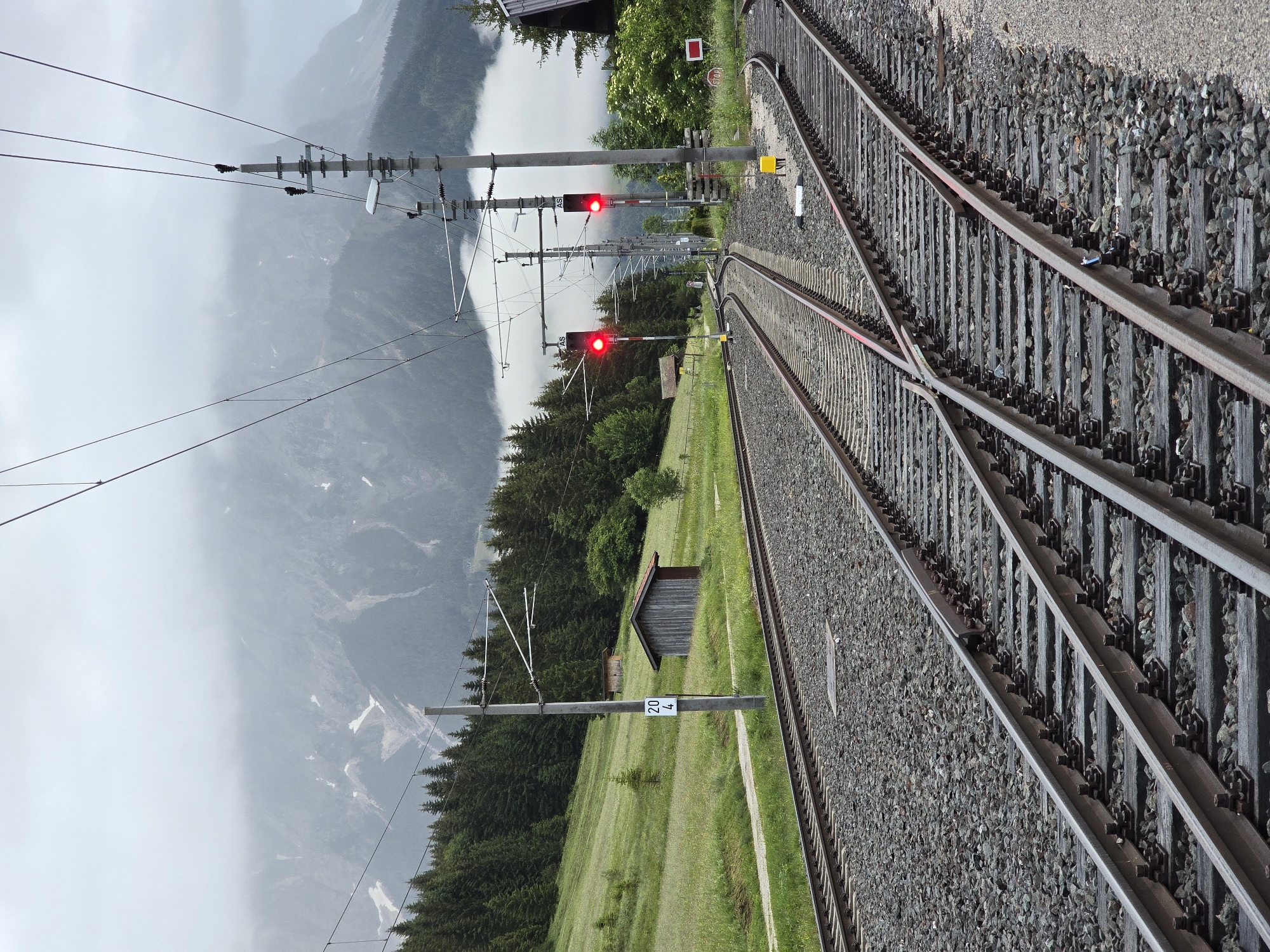 The image depicts a railway junction in Lermoos, Austria, on a cloudy day. Twin sets of parallel railway tracks dominate the foreground, converging and splitting as they recede into the distance. The tracks are laid on a ballast of gray stones and supported by dark wooden sleepers. Overhead, a network of electrical wires for the train system stretches across the frame, suspended by utilitarian poles.
Two railway signals are visible, both displaying a red light, indicating a stop. The signals are mounted on poles, one on the left and another on the right side of the tracks further down the line. A small, weathered wooden shed is situated on a grassy embankment to the left of the tracks, and beyond it, a dense forest of dark green pine trees covers the hillside.
In the background, the imposing silhouette of a mountain range looms, its peaks partially obscured by a thick layer of fog and clouds. Patches of snow are visible on the mountain slopes, suggesting a higher elevation and cooler climate. The overall environment is rural and mountainous, characteristic of the Austrian Alps. There are no people visible in the image.