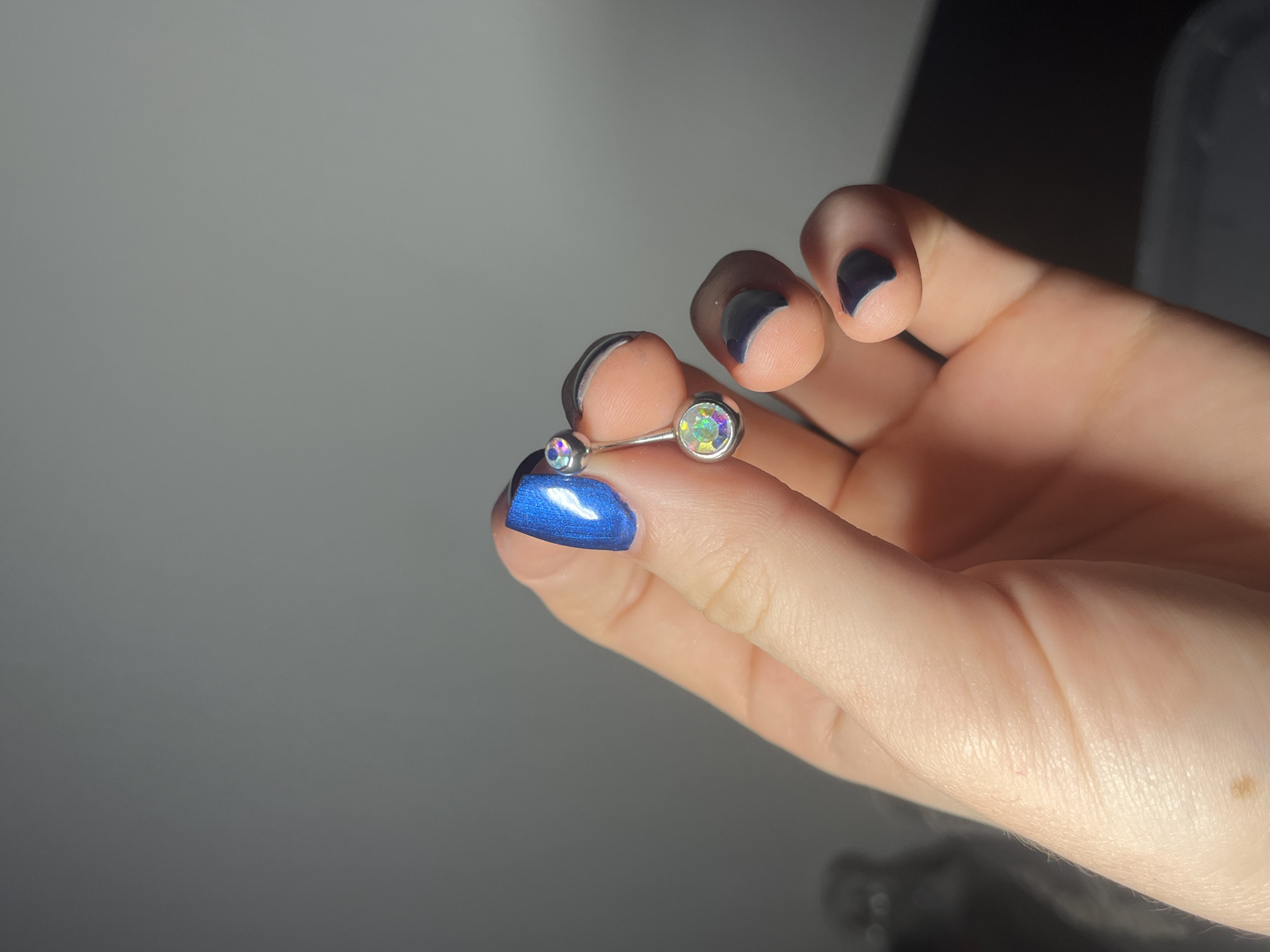 A fair-skinned hand holds a curved barbell piercing with two iridescent gems in a well-lit environment. The index finger nail is painted blue, while the other visible fingernails are painted dark gray or black. A single mole is visible on the palm. The background appears to be a solid, neutral-toned surface.