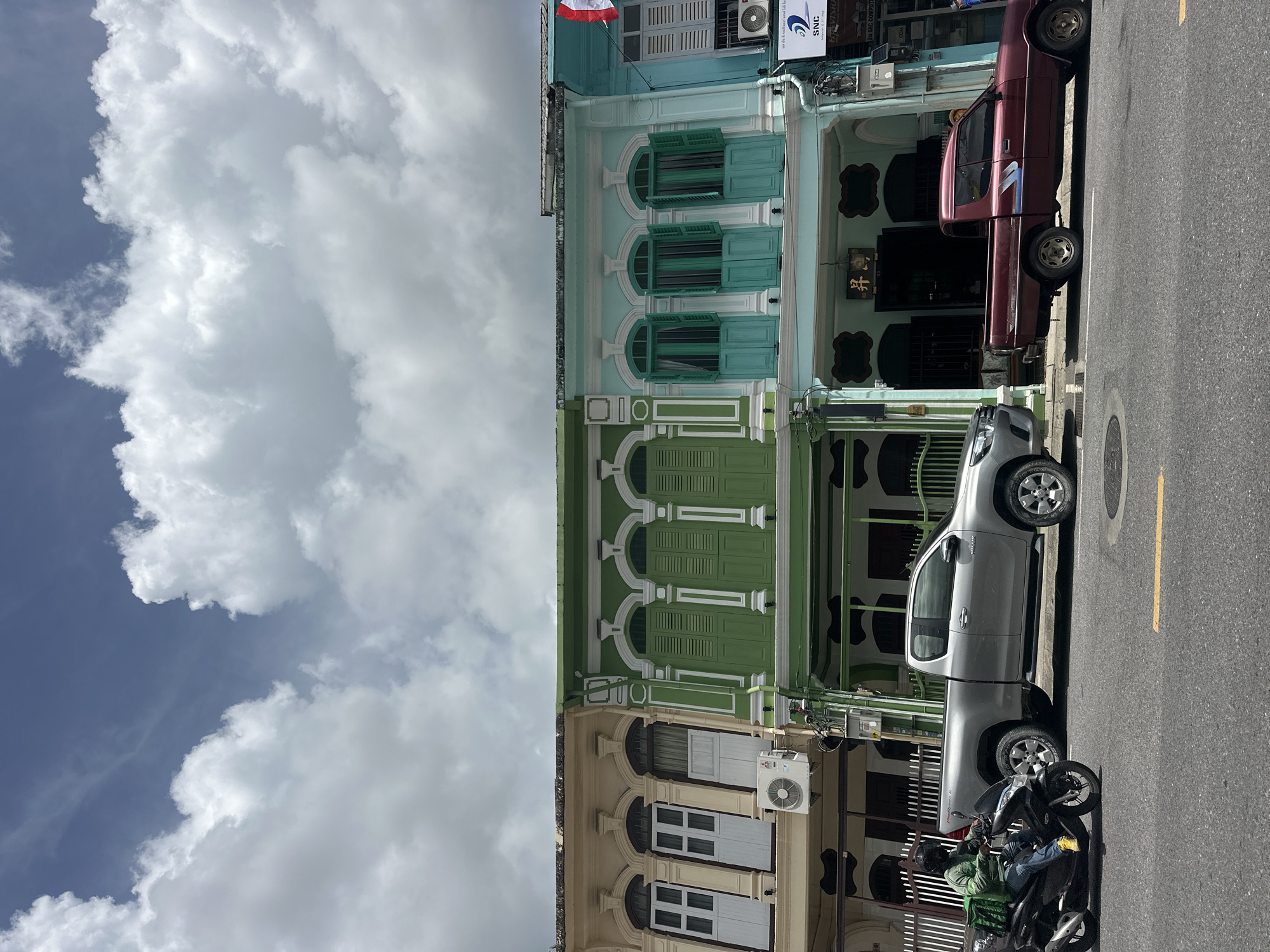 This is a wide shot of a street scene in Phuket City Municipality, Thailand, focusing on a row of colorful shophouses. The sky above is partially cloudy with a mix of white cumulus clouds against a blue background.
In the foreground, a gray pickup truck is parked on the right side of the road. To its left, another silver pickup truck is parked, and a person wearing a green jacket and blue jeans is riding a black motorcycle with a green delivery bag on the back. The motorcycle is moving from left to right. A yellow line is visible on the asphalt road, indicating a lane marker.
The row of shophouses features a blend of architectural styles, with ornate details and varying pastel colors. From left to right, the first shophouse is a pale yellow with white-trimmed arched windows and shutters. An outdoor air conditioning unit is visible on its facade. The next shophouse is painted in a vibrant green, also with arched windows and shutters. The third shophouse is a light turquoise with green shutters. A sign with Chinese characters is mounted above the entrance of this shophouse. The fourth shophouse, partially visible on the far right, is a bright blue. A Thai flag, red and white striped, is attached to the top of this building.
The buildings appear to be two stories high, with the upper floors featuring the distinctive arched windows and decorative elements. Electrical wires and conduits are visible along the sides of the buildings. The street appears to be a commercial area with shops or businesses located within the shophouses. The overall impression is a bright, urban street scene with elements of traditional architecture.