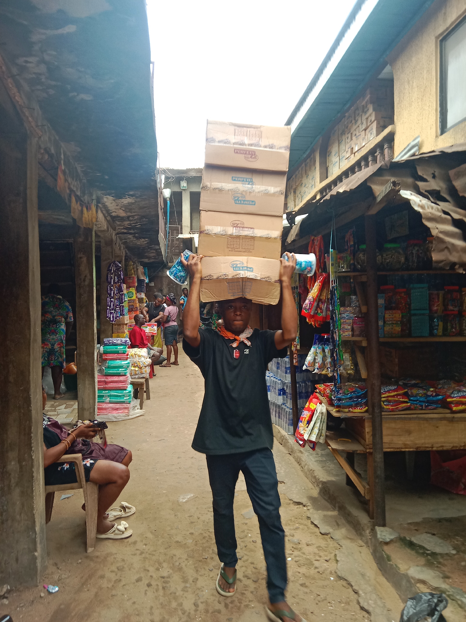 In Onitsha, Nigeria, a young man walks down an outdoor market lane carrying a stack of approximately six cardboard boxes on his head. He wears a black t-shirt, dark pants, and flip-flops. He also has an orange scarf or bandana around his neck. Other people are visible in the background of the lane, including vendors at stalls selling various goods, and other individuals walking and sitting. The lane is narrow and enclosed by buildings and stalls on either side. The ground appears to be uneven and made of dirt or concrete. Merchandise, snacks, and drinks are on display at stalls and on the right, behind the man.