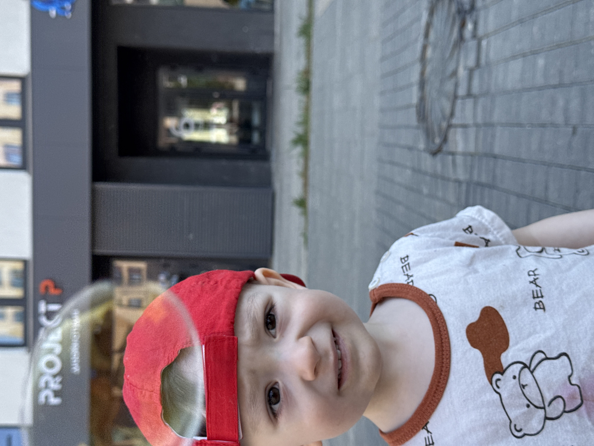 A young child wearing a red cap and a white shirt with a bear design is looking at the camera. The child is standing on a sidewalk with gray paving stones. Behind the child, a building with a sign that reads "PROJECT P" is visible. The reflection in the building's window reveals more of the city environment. The scene appears to be in Izhevsk, Russia, based on the Cyrillic script used in the "PROJECT P" sign.