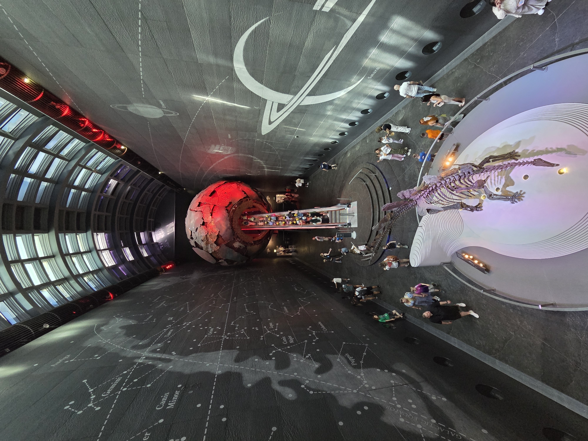Here's a detailed description of the image:
The image presents an unusual, overhead perspective of a large indoor space, likely a museum hall. The room is dominated by a dark, textured floor with constellations and galactic equator markings. On the left, a curved wall features rows of window-like structures with bright light shining through. Towards the center, a large, spherical object, possibly a globe or planet model, glows with red light.
To the right of the sphere is a raised platform displaying a dinosaur skeleton. Several people are scattered throughout the scene, observing the exhibits. The ceiling features planetary designs, including the planet Saturn and its rings. The location is likely the Natural History Museum in London, United Kingdom.