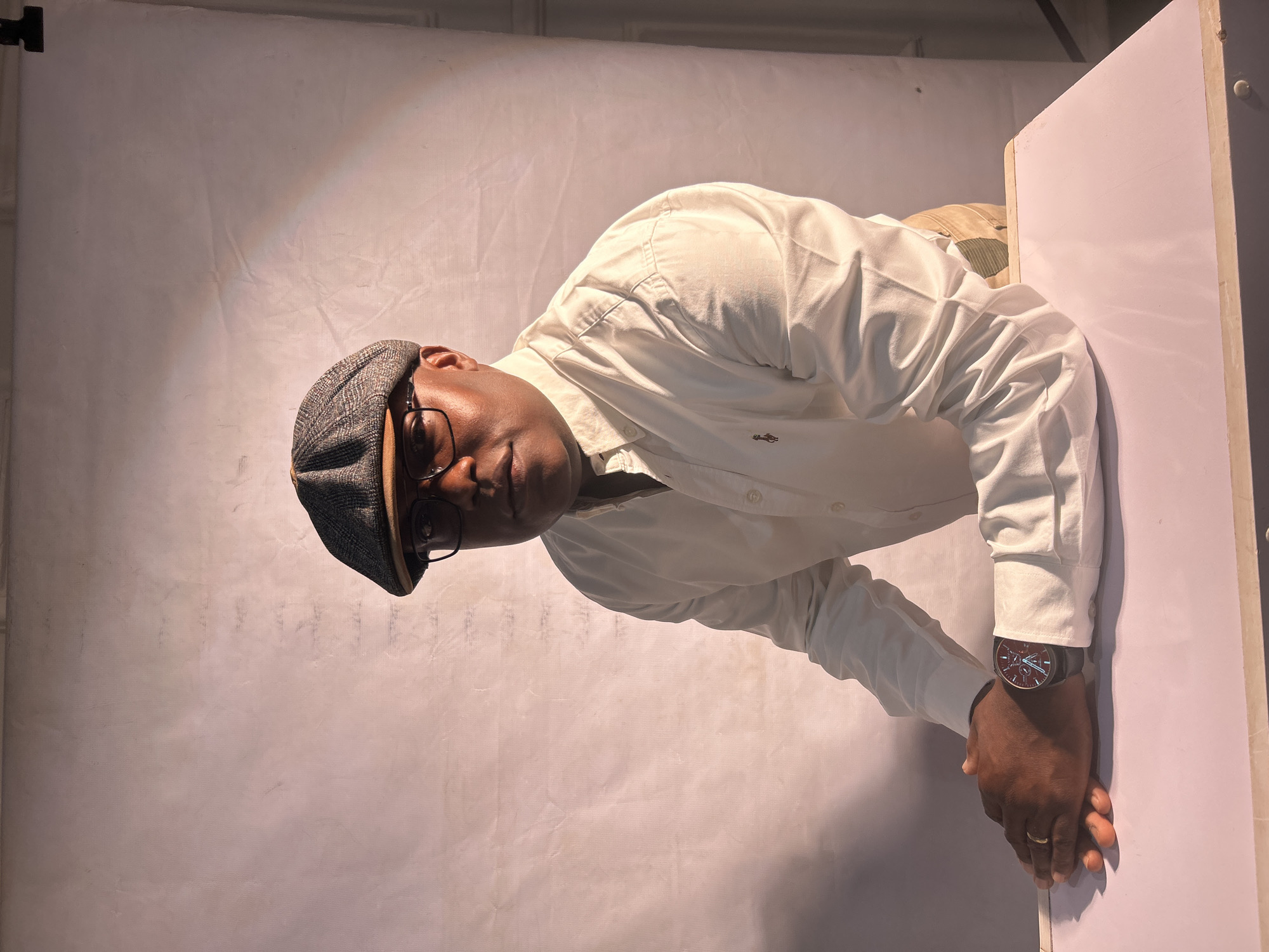A man wearing a grey newsboy cap and glasses, and a white long-sleeved shirt, stands in front of a white backdrop. His hands, adorned with a wristwatch on his left wrist, rest on a white surface. The location is presumed to be a photography studio in Jos, Nigeria.