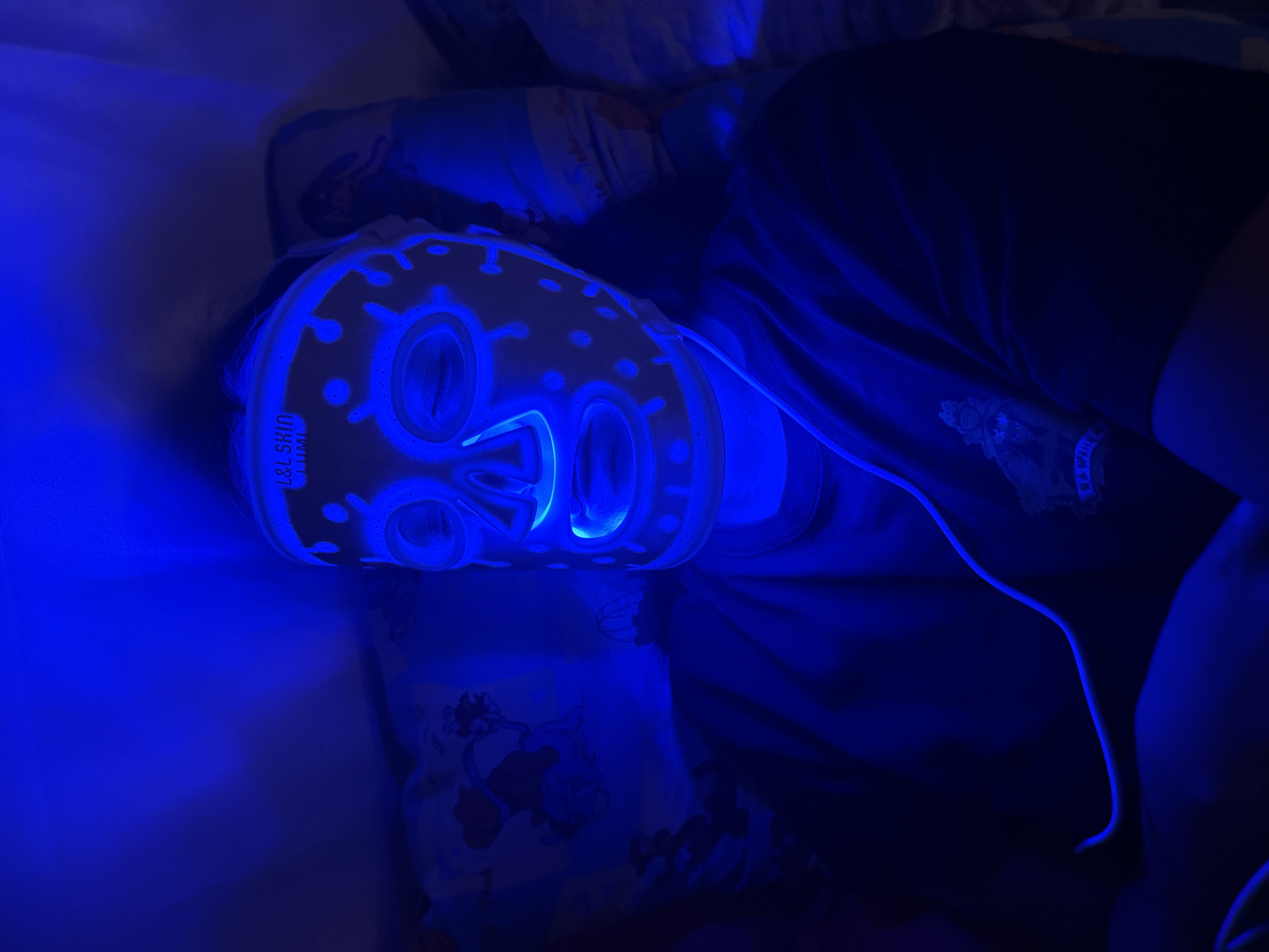 A person lies supine in an interior setting in Красный Маяк, Russia. The person wears a blue LED facial mask. The environment includes bedding, a wire from the mask, and a dark t-shirt.