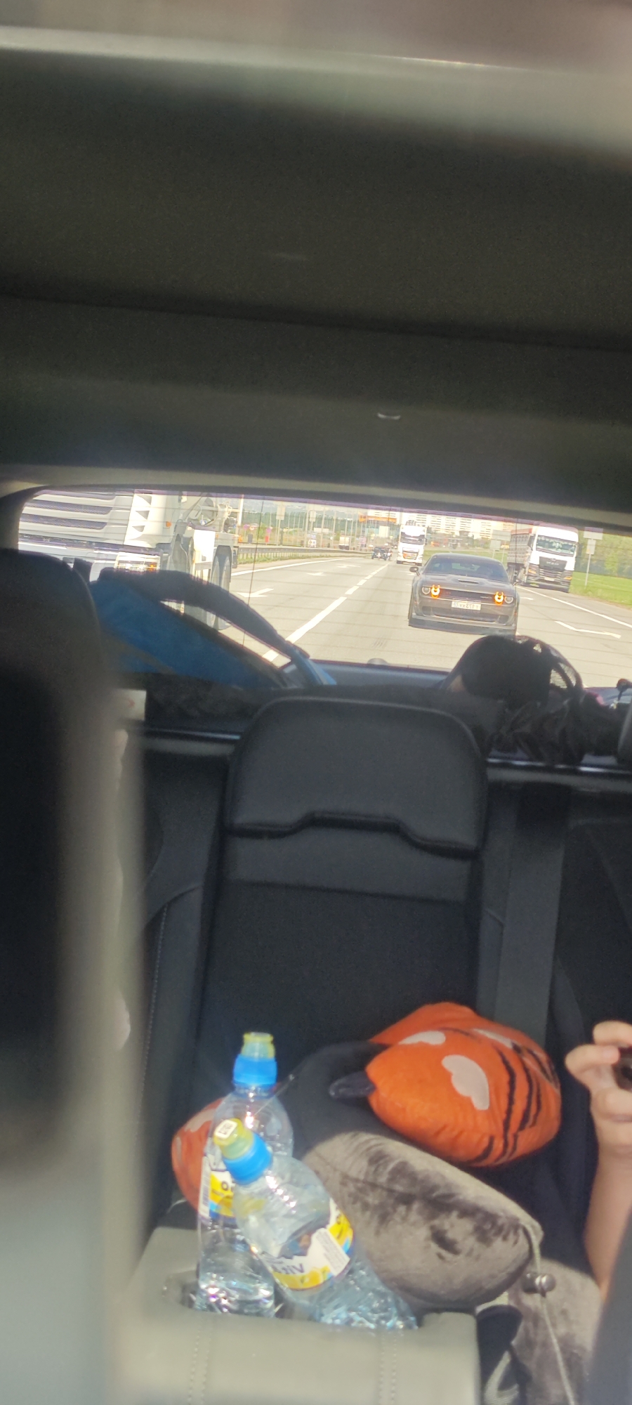 The image depicts a view from inside a car, looking out the rear window onto a highway. The scene appears to be taken during daytime. Multiple vehicles including trucks and cars are visible on the highway. In the foreground, the back seat of the car is visible with bottles of water and a stuffed animal. The license plate of the car driving in front has the code "63" indicating this is likely in the city of Prigorodny, Russia.