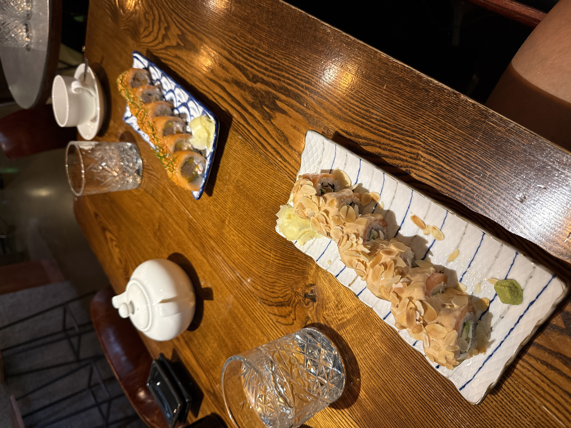 The image depicts a wooden table in a dimly lit restaurant, likely in Saint Petersburg, Russia, given the presence of a white teapot, a common item in Russian tea culture. On the table, two plates of sushi are arranged. The plate in the foreground holds sushi rolls topped with sliced almonds and a creamy sauce, accompanied by pickled ginger and a dollop of wasabi. In the background, another plate showcases sushi rolls coated in orange roe. Nearby, a glass of water, a teacup and saucer, and a small black dish for soy sauce are visible. The restaurant appears to have a modern, somewhat rustic aesthetic, with glimpses of chairs and other tables in the background. The lighting suggests an evening setting.